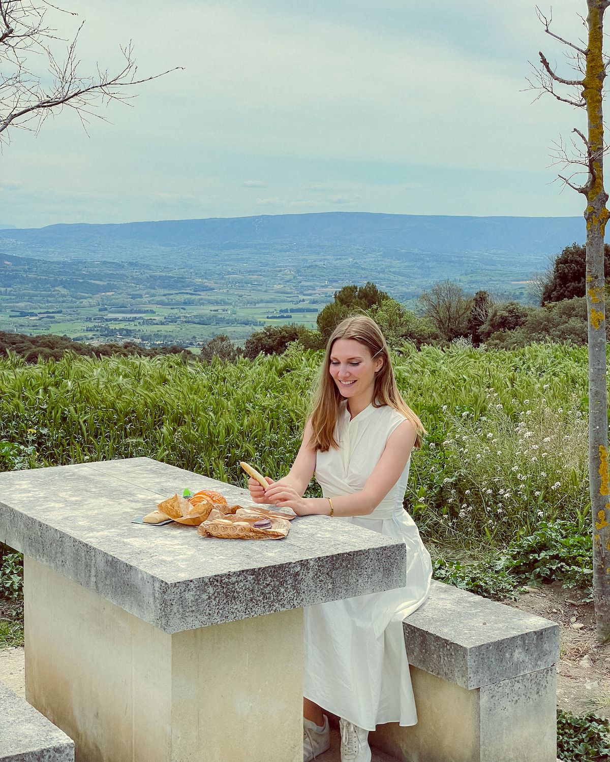 Blonde girl in white dress having a picnic on a concrete picnic table overlooking the grassy and hilly countryside of Provence