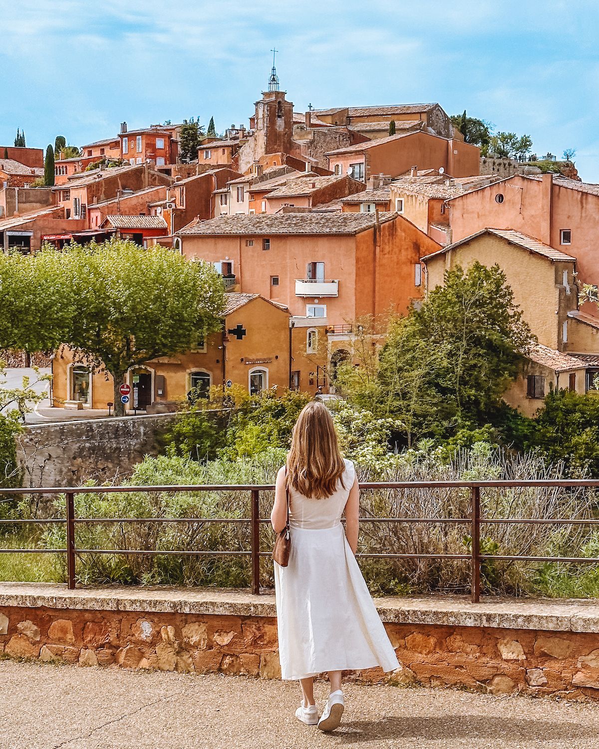 Blonde girl in white dress looking at ochre coloured buildings in the village of Roussillon