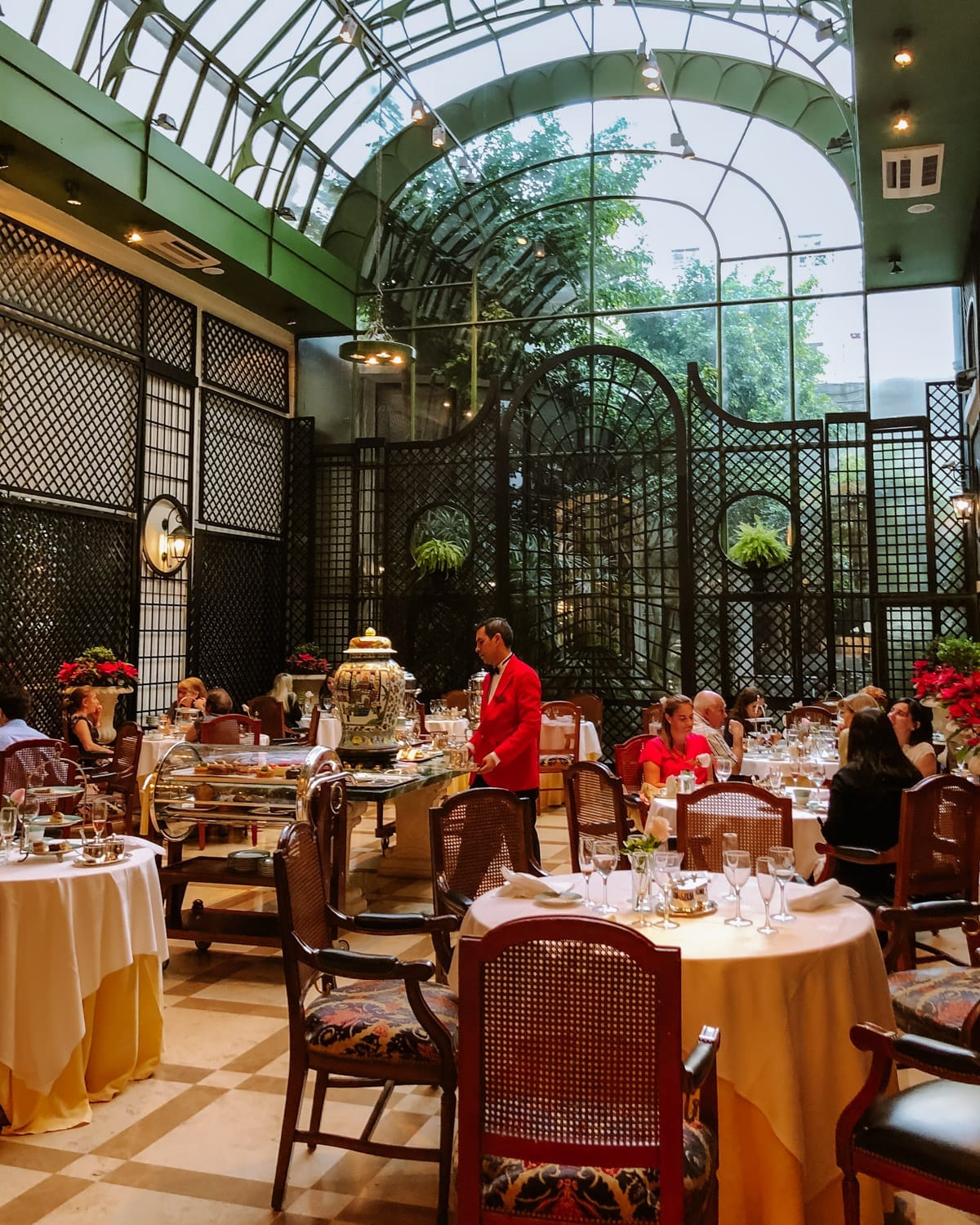 Elegant glass atrium with guests dining on white-clothed tables as a server in red offers tea service at L’Orangerie.