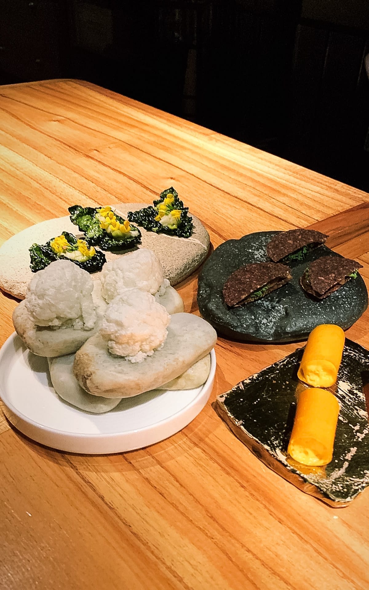 An artful selection of small bites served on textured stones, featuring foams, crisp leaves, and golden mousse rolls.