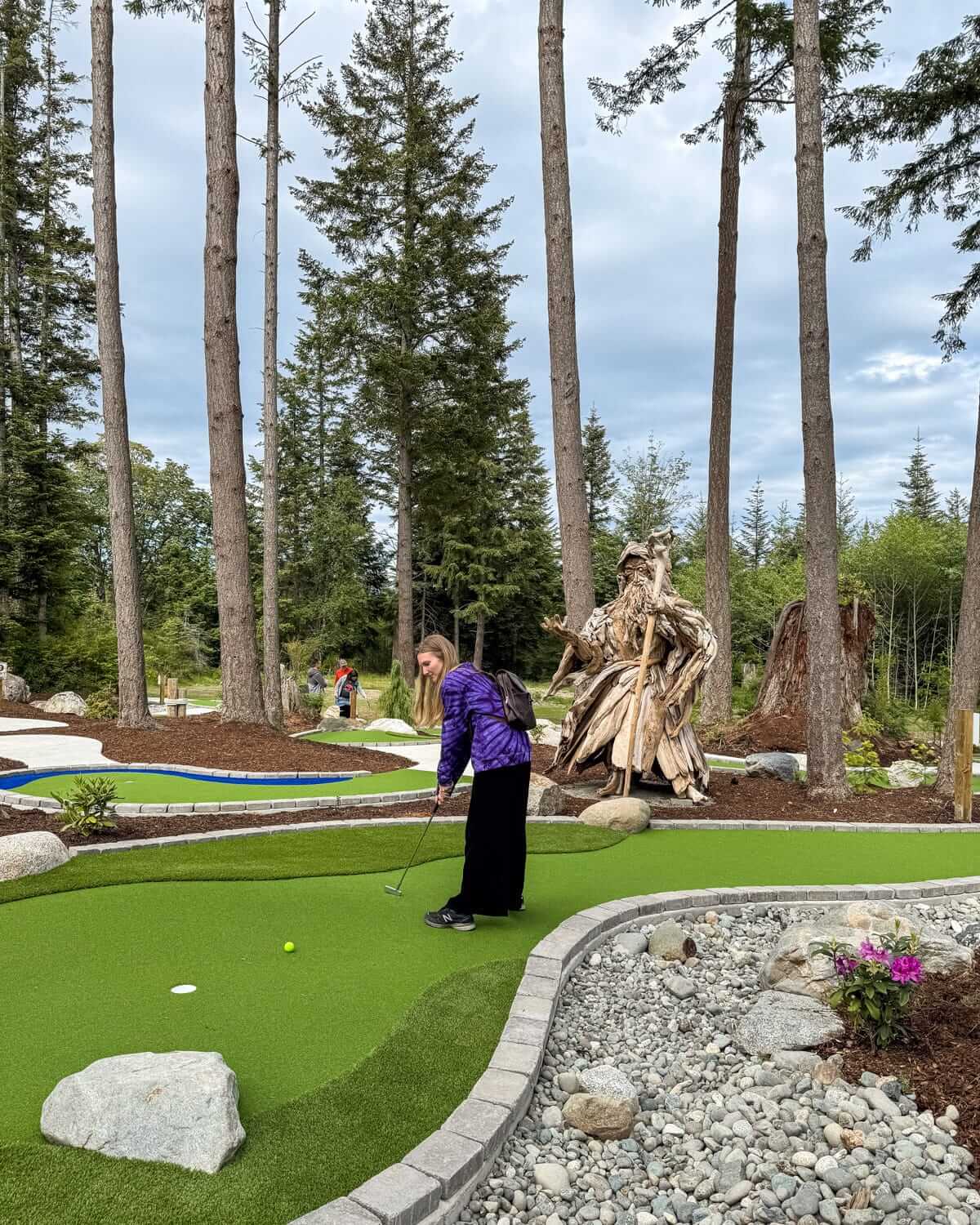 Cec playing mini-golf on a well-manicured green course at Quadra Island Golf Course surrounded by tall trees and whimsical driftwood sculptures.