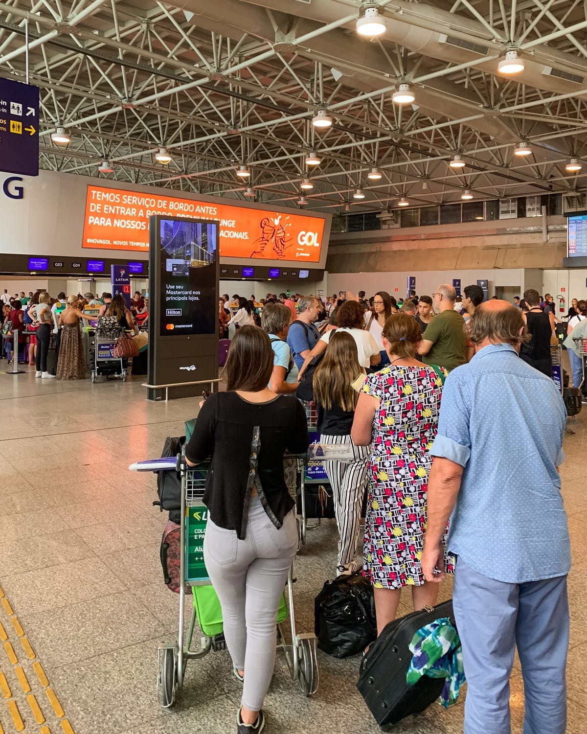 A long, chaotic queue of travellers at Rio’s international airport check-in area under bright lights and industrial architecture.