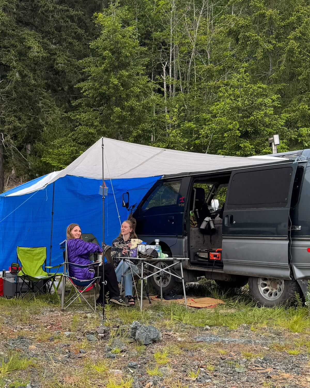 Cec and a friend relax in camp chairs under a tarp beside a Delica camper van on Quadra Island.