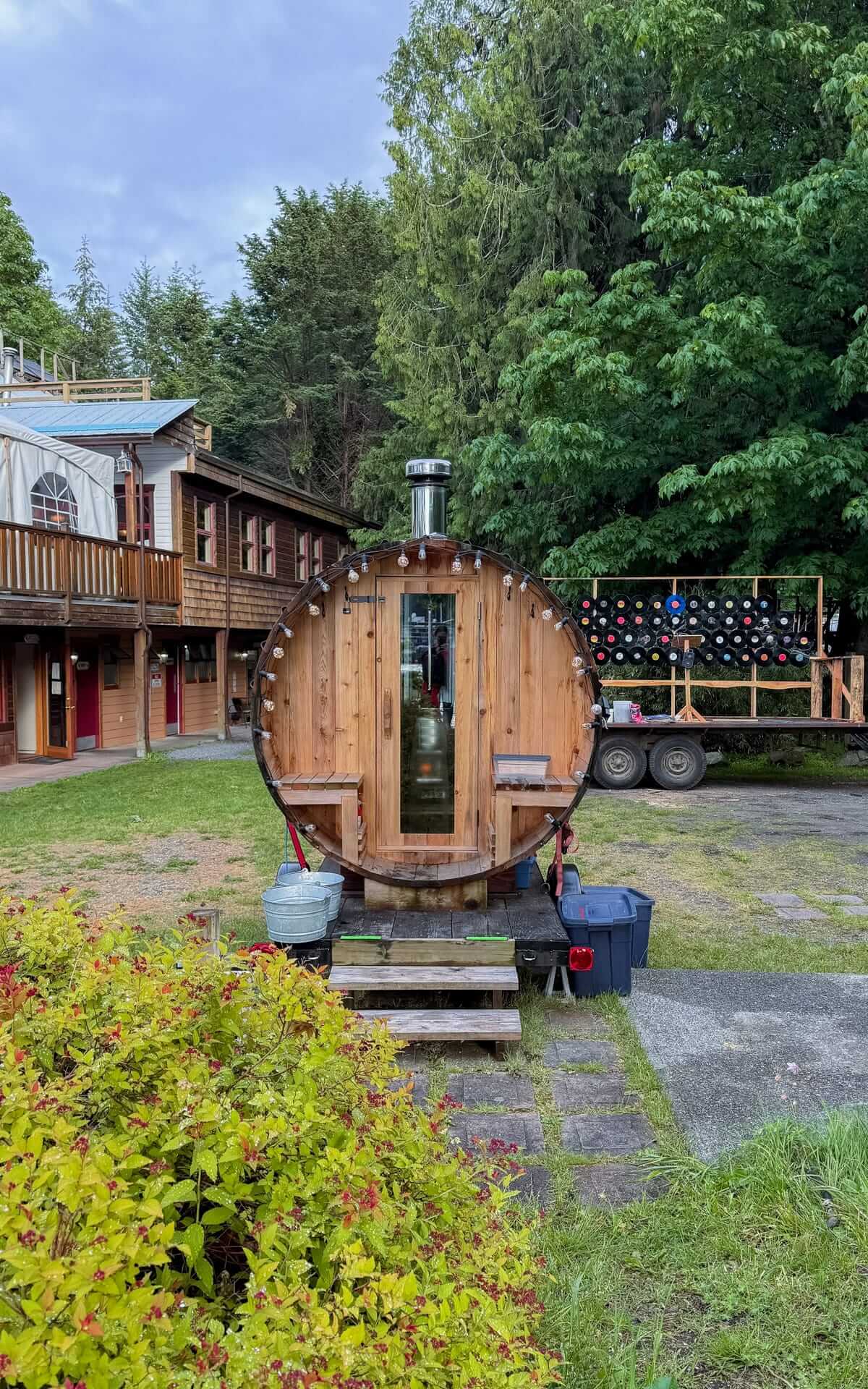 A barrel-shaped sauna with fairy lights sits in a grassy yard at Heriot Bay Inn, backed by a rustic lodge and surrounded by lush trees.