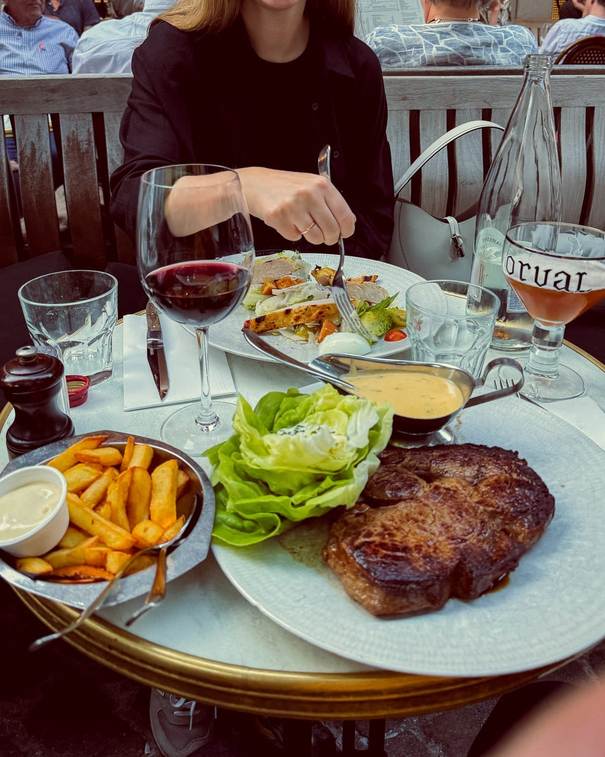 A juicy grilled steak, Belgian fries with mayo, chicken caesar salad, beer and red wine rest on a bistro table at Bourla in Antwerp.
