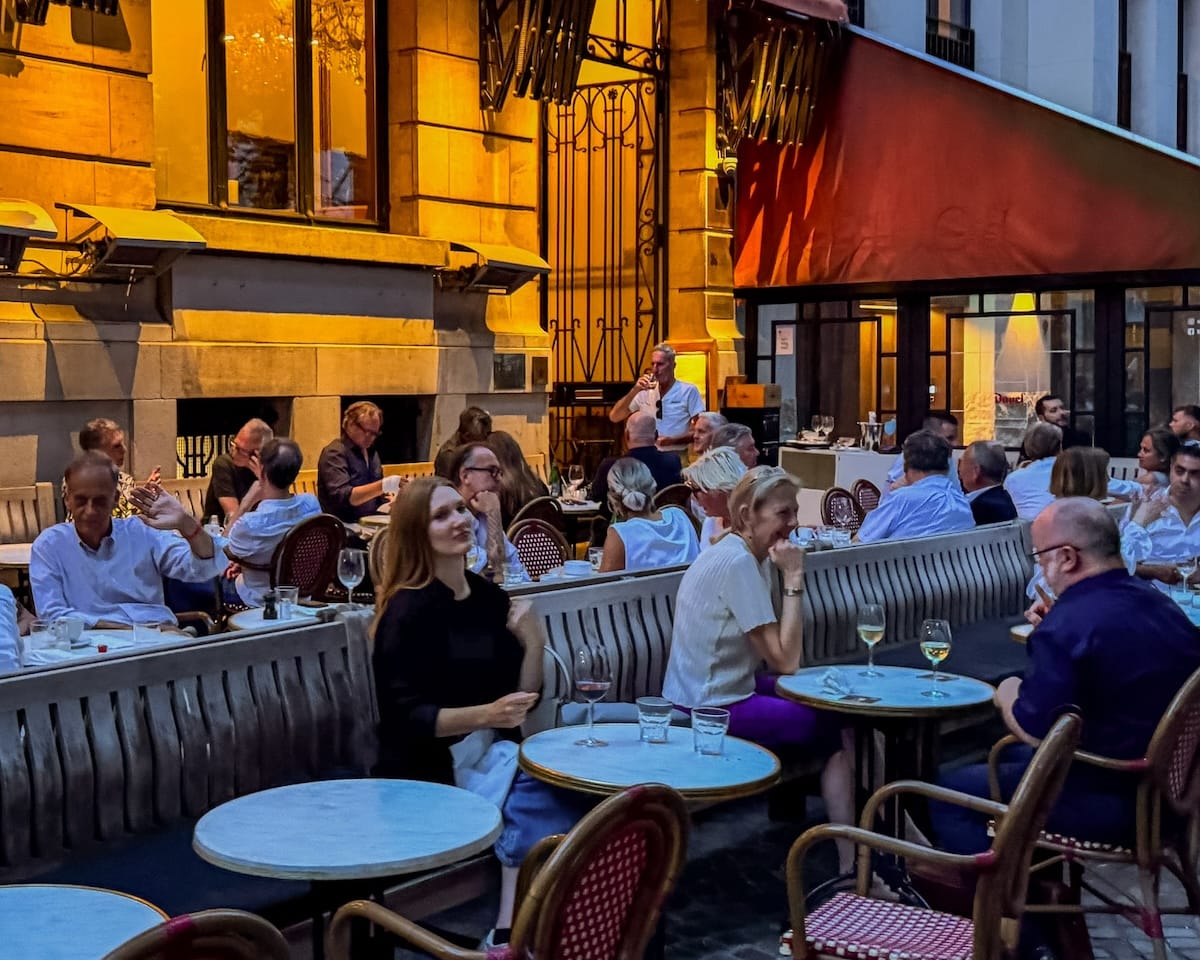 People dining al fresco on the chic terrace of Bourla in Antwerp at dusk, with warm yellow lighting and red and white French bistro chairs.