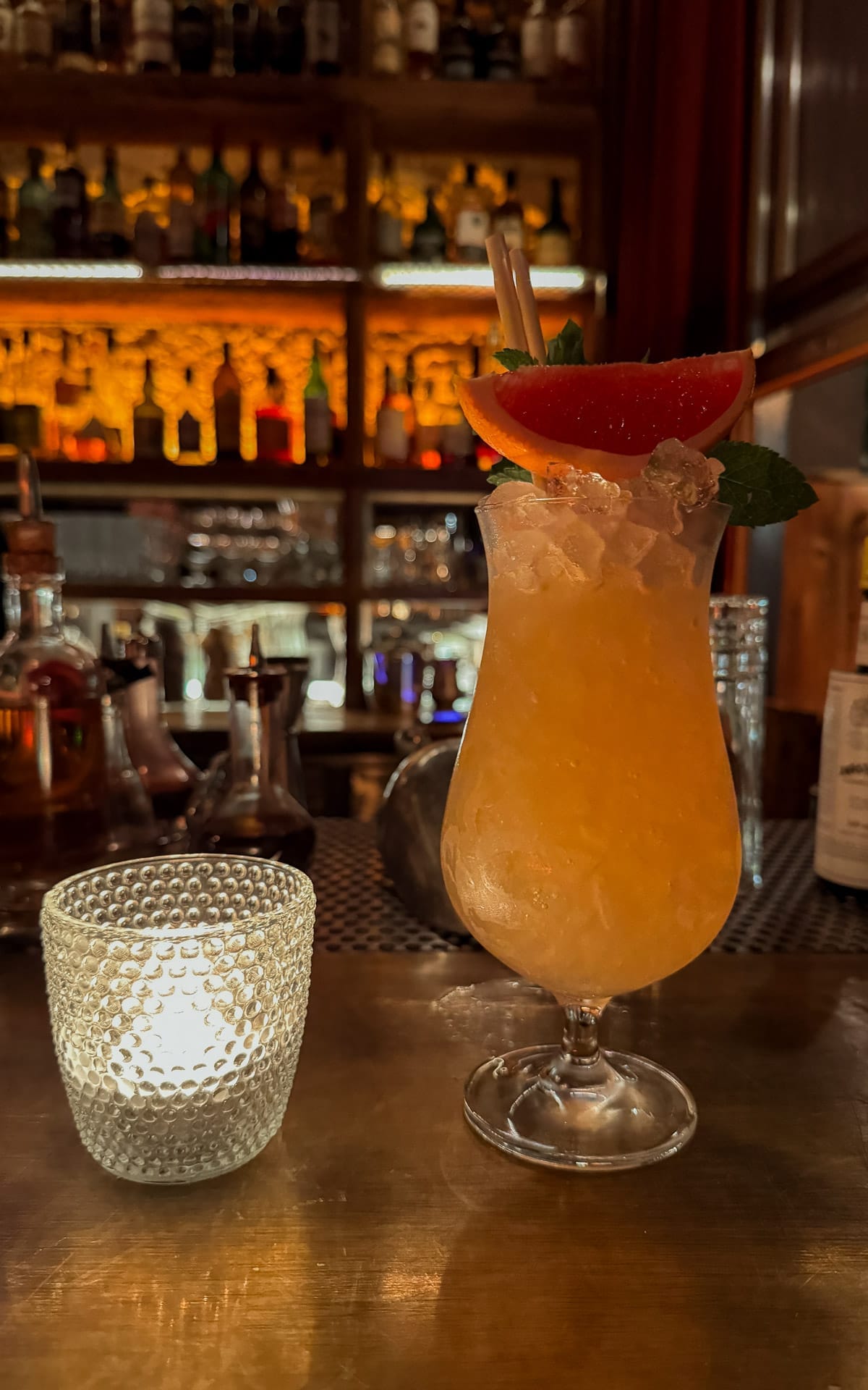 A fruit punch cocktail garnished with mint and grapefruit and a candle rest on the bar of Dogma, with shelves of spirits in soft focus.