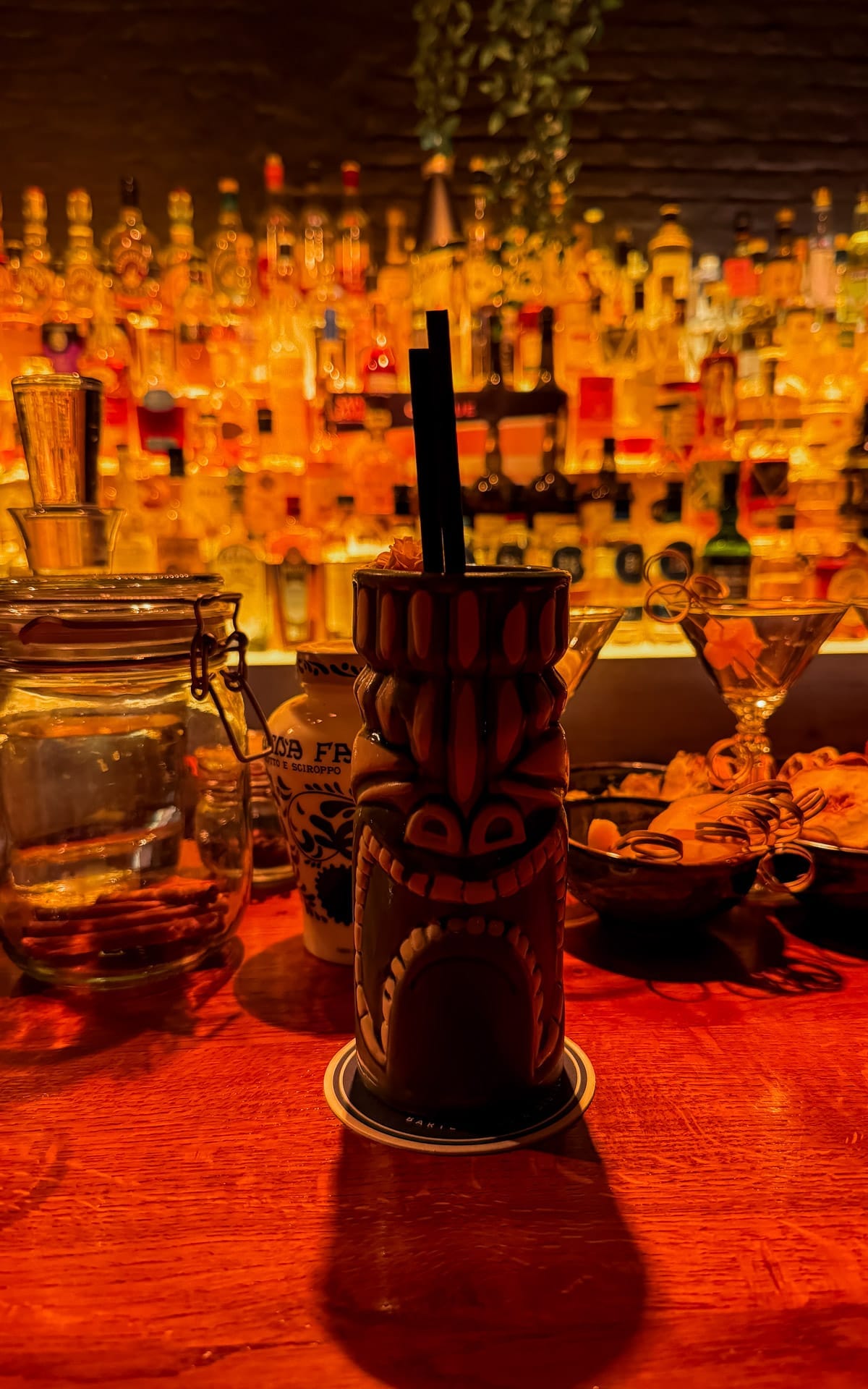 A tiki mug with a straw sits at a dimly lit bar surrounded by bottles and bar tools, glowing in a warm amber light at Belroy's Bijou.