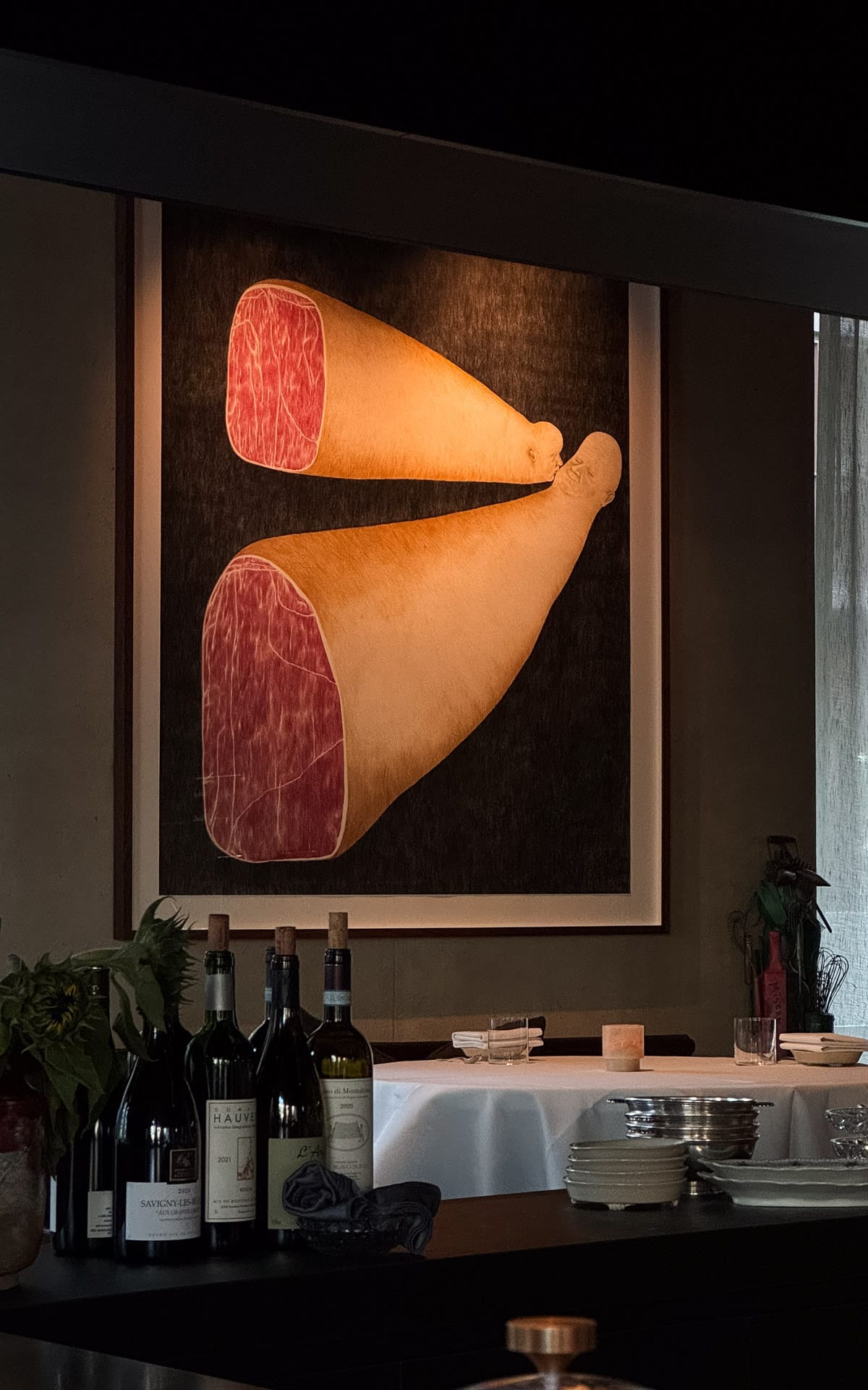 Close-up of a framed contemporary painting of two ham legs above a counter with wine bottles in Misera's chic dining space.