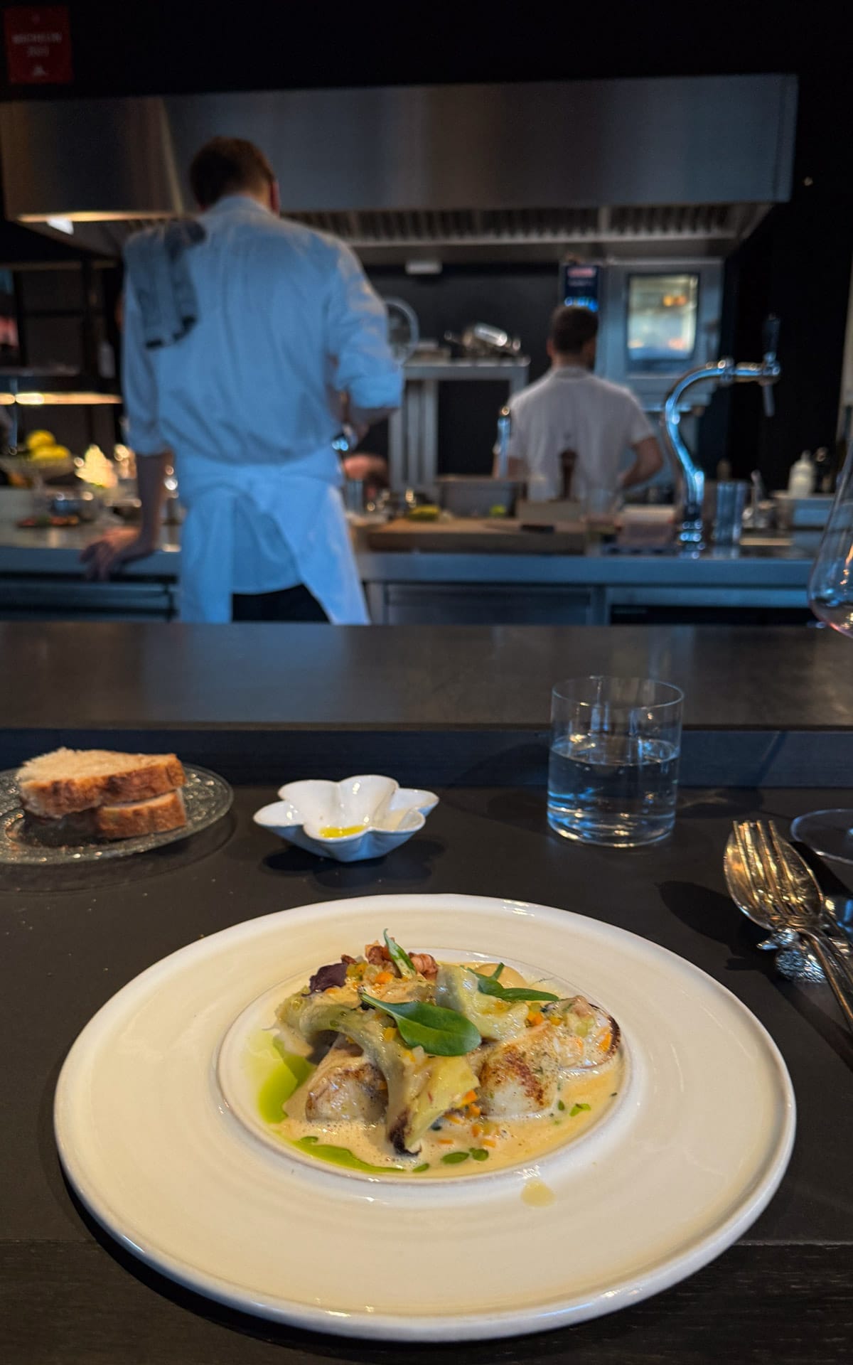 A beautifully plated dish of white fish is served at a sleek black counter, with chefs working in the open kitchen in the background at Misera..