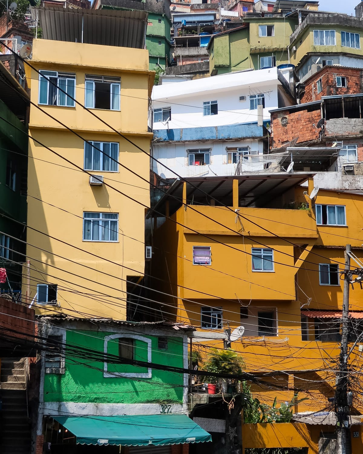 Tall, closely packed buildings painted in bold colours like green, yellow, and orange, connected by a web of visible wiring in Rocinha favela.