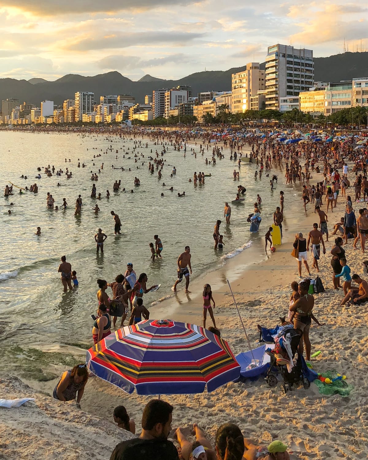 A packed Ipanema Beach filled with locals and tourists enjoying the water as the sun sets over the mountains in the background.