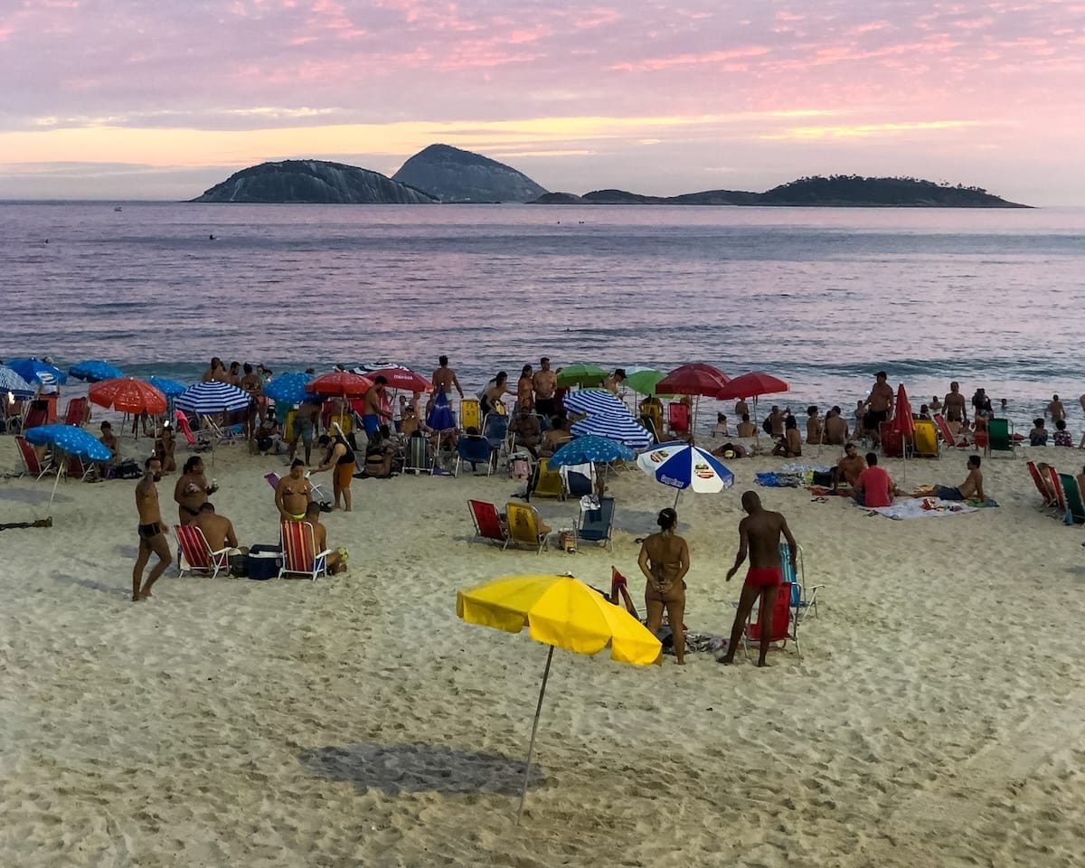 Colourful umbrellas and beachgoers dot the sand at Ipanema Beach during sunset, with pink and purple skies and distant rocky islands.