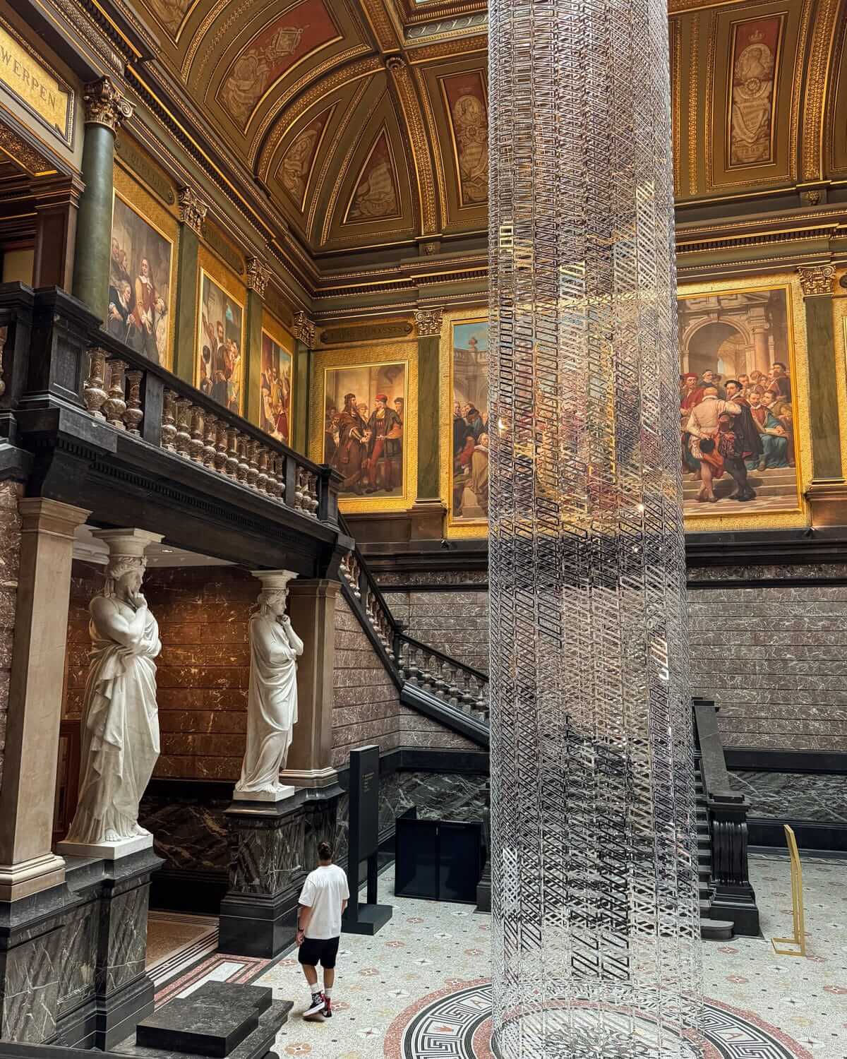 Grand museum hall of KMSKA with classical statues, arched staircases, artwork adorned walls and ceilings, and tall transparent sculpture installation.