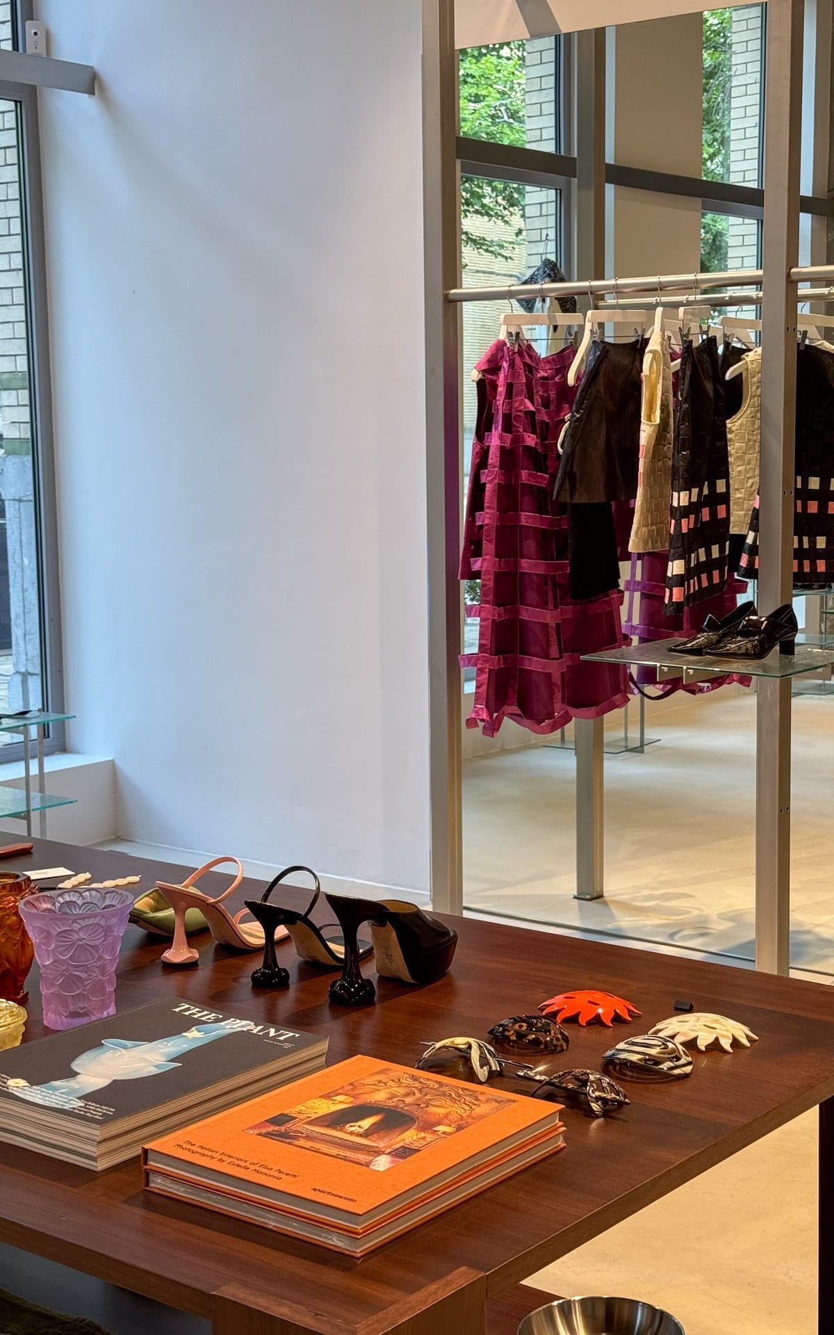 Curated fashion and design items, including statement heels, clothing, books, sunglasses, and hair pins displayed on a wooden table at The Menu in Antwerp.