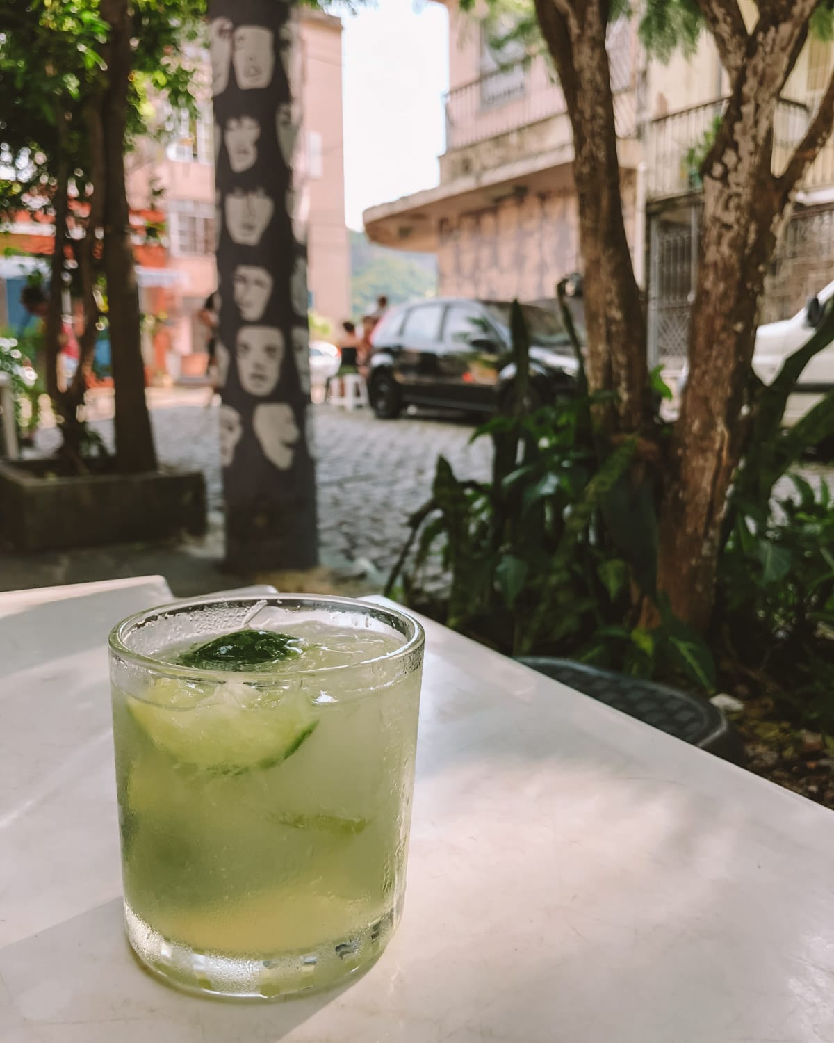Refreshing glass of caipirinha with lime and ice on a white table, shaded by trees and surrounded by a quiet cobbled street scene in Santa Teresa..