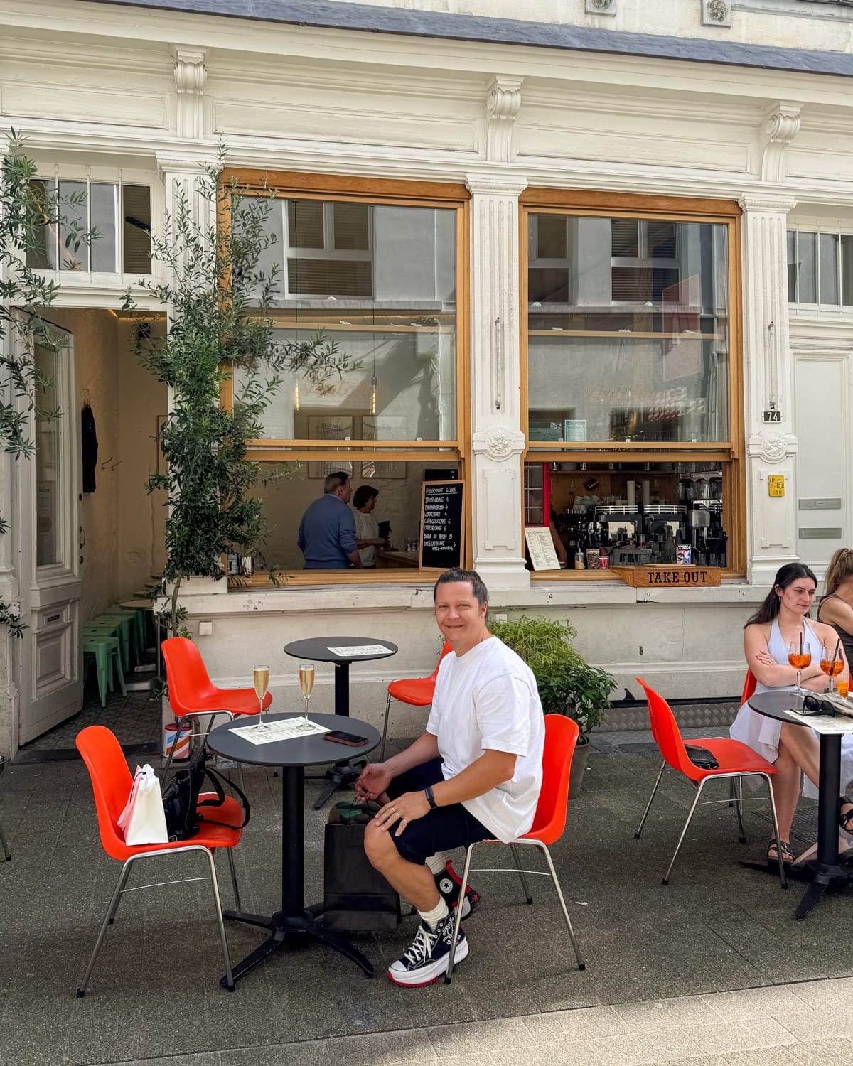 Ari smiling at a table outside a Antoine cafe with Aperol-orange chairs, champagne glasses, and people relaxing in warm weather.