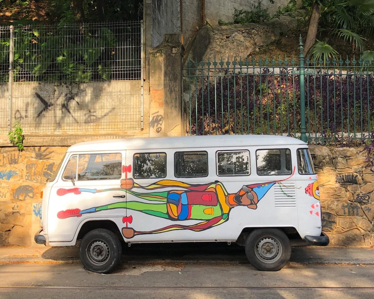 White van parked on a city street in Santa Teresa, painted with a colourful abstract figure wearing a party hat, against a graffiti wall.
