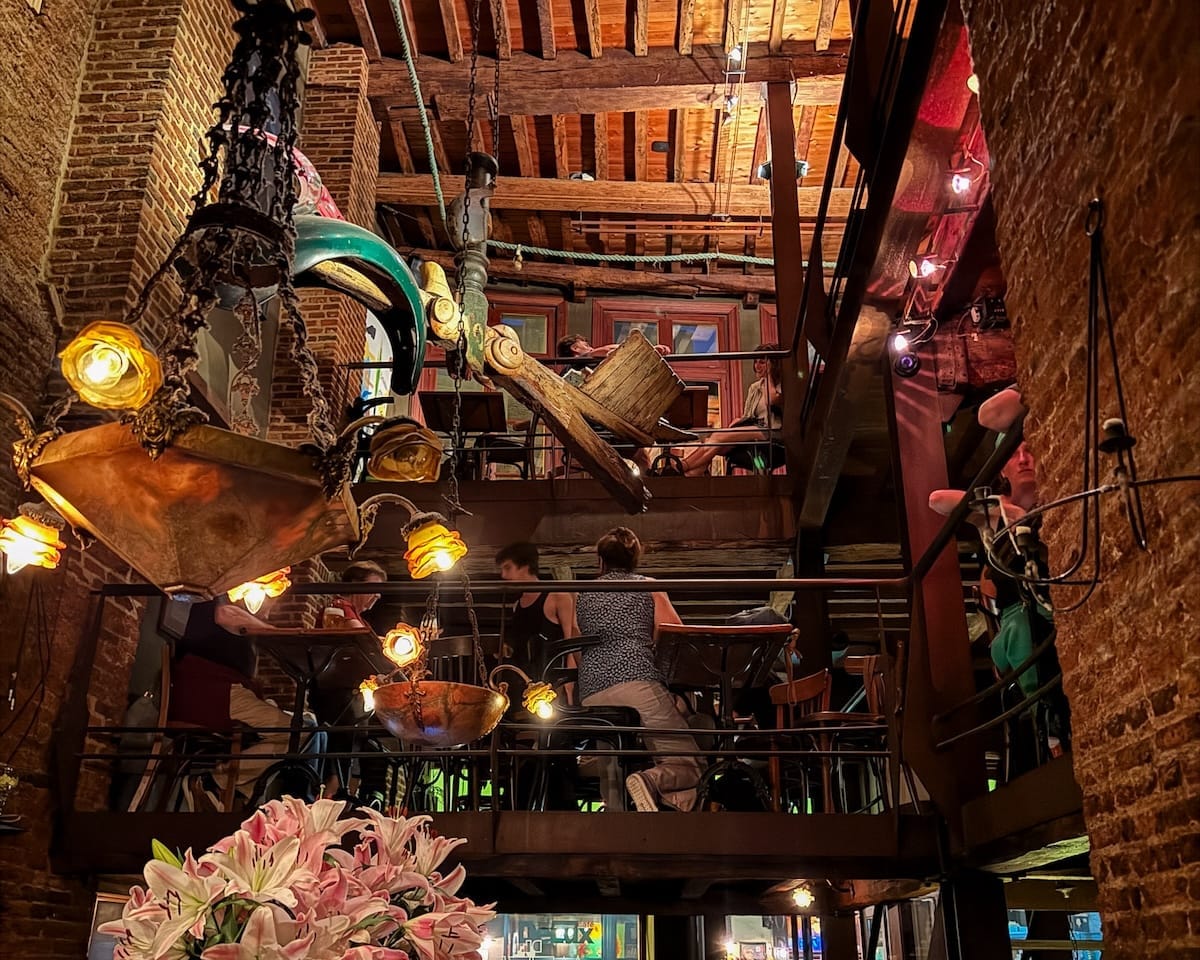 Warmly lit bar with rustic wood beams, hanging lamps, and guests dining on upper and lower levels in the unique multi-tier space of De Muze Cafe.