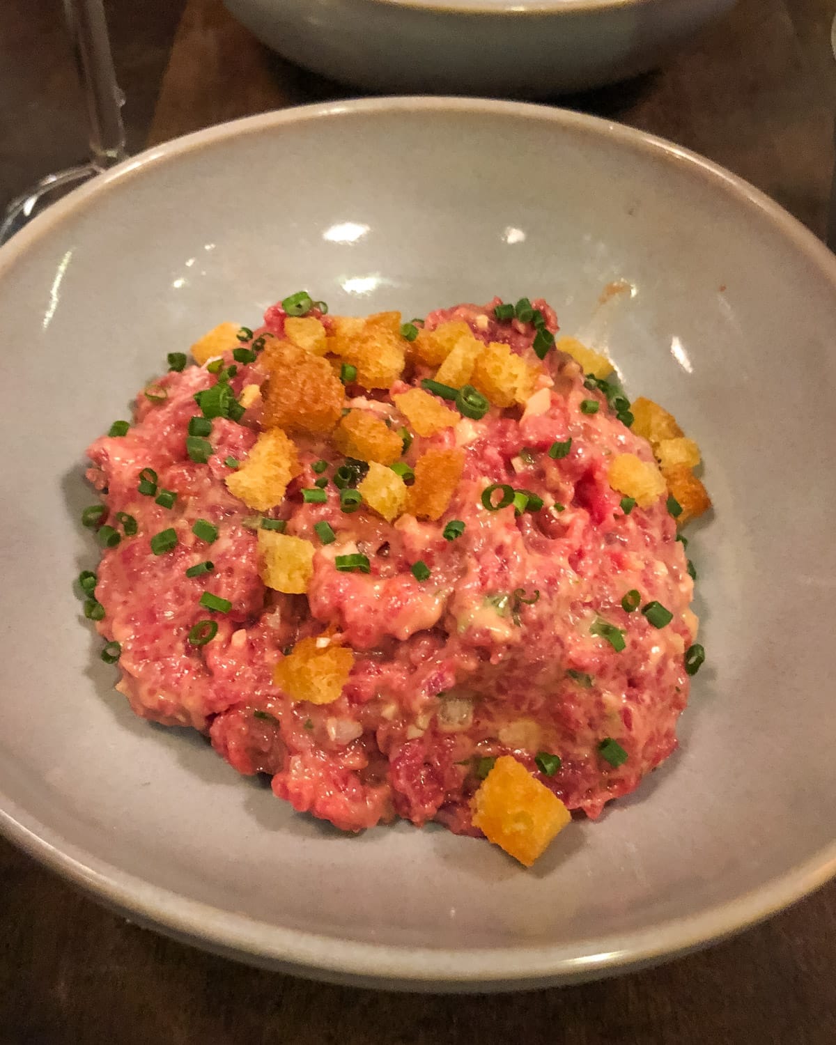 Close-up of a dish of beef tartare topped with chopped chives and golden croutons in a gray ceramic bowl at a CT Boucherie in Rio.