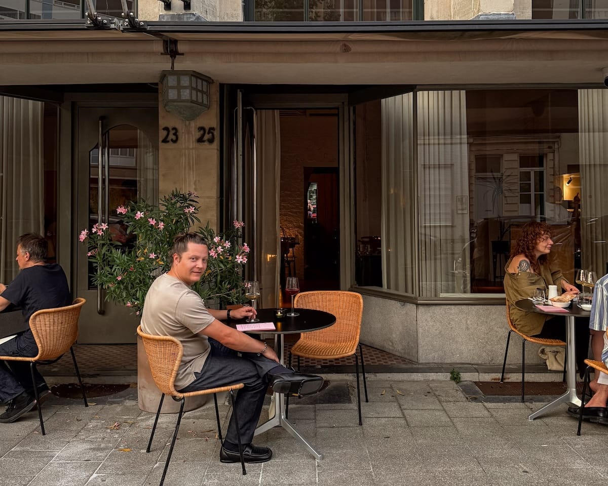 Ari enjoys an evening aperitif at a cozy sidewalk table outside Le John, surrounded by relaxed guests.