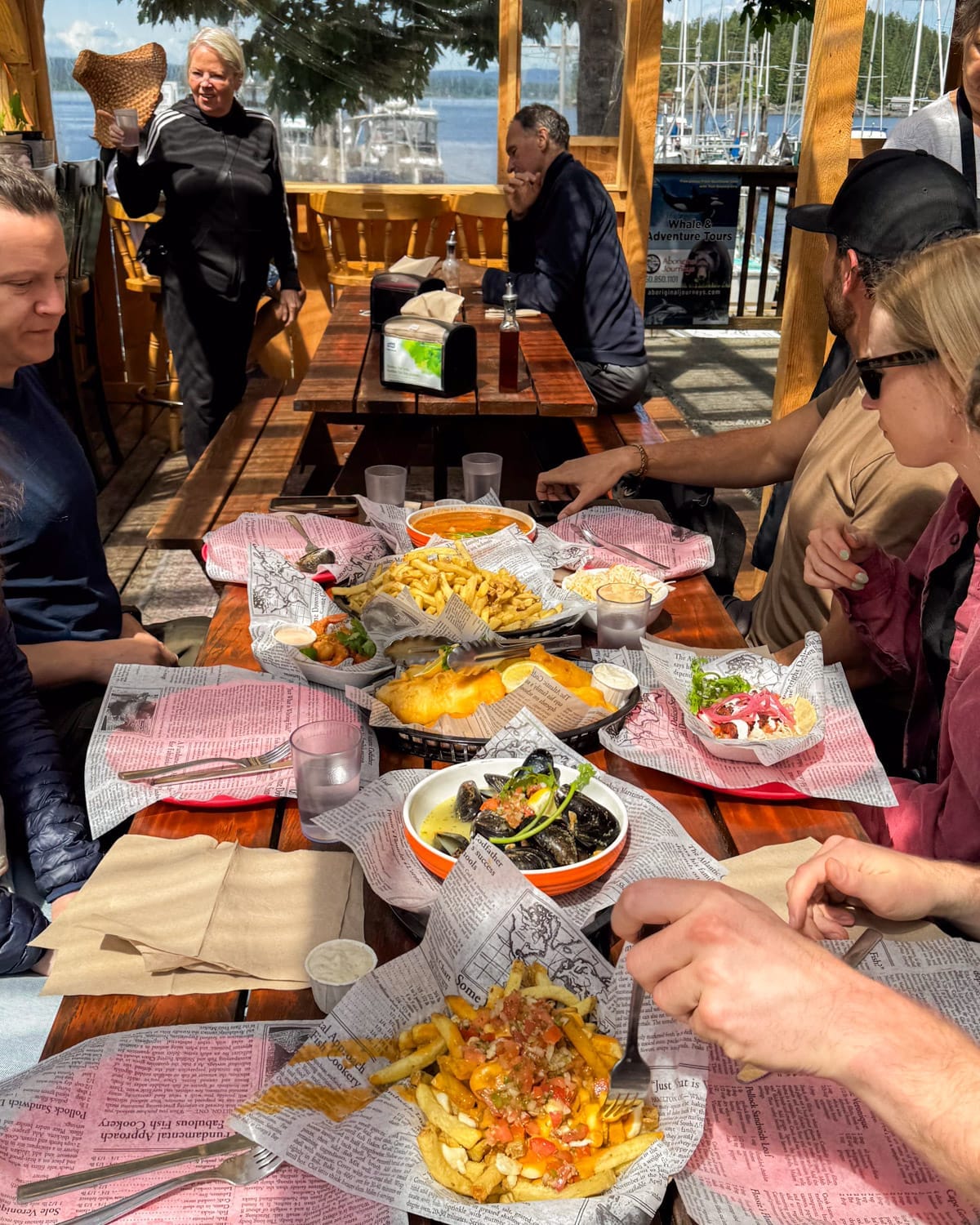 Outdoor seafood feast on a wooden patio overlooking a marina at Coveside Seafood Eatery, with a table full of fries, mussels, tacos, and fish & chips - one of the best places to eat on Quadra Island.