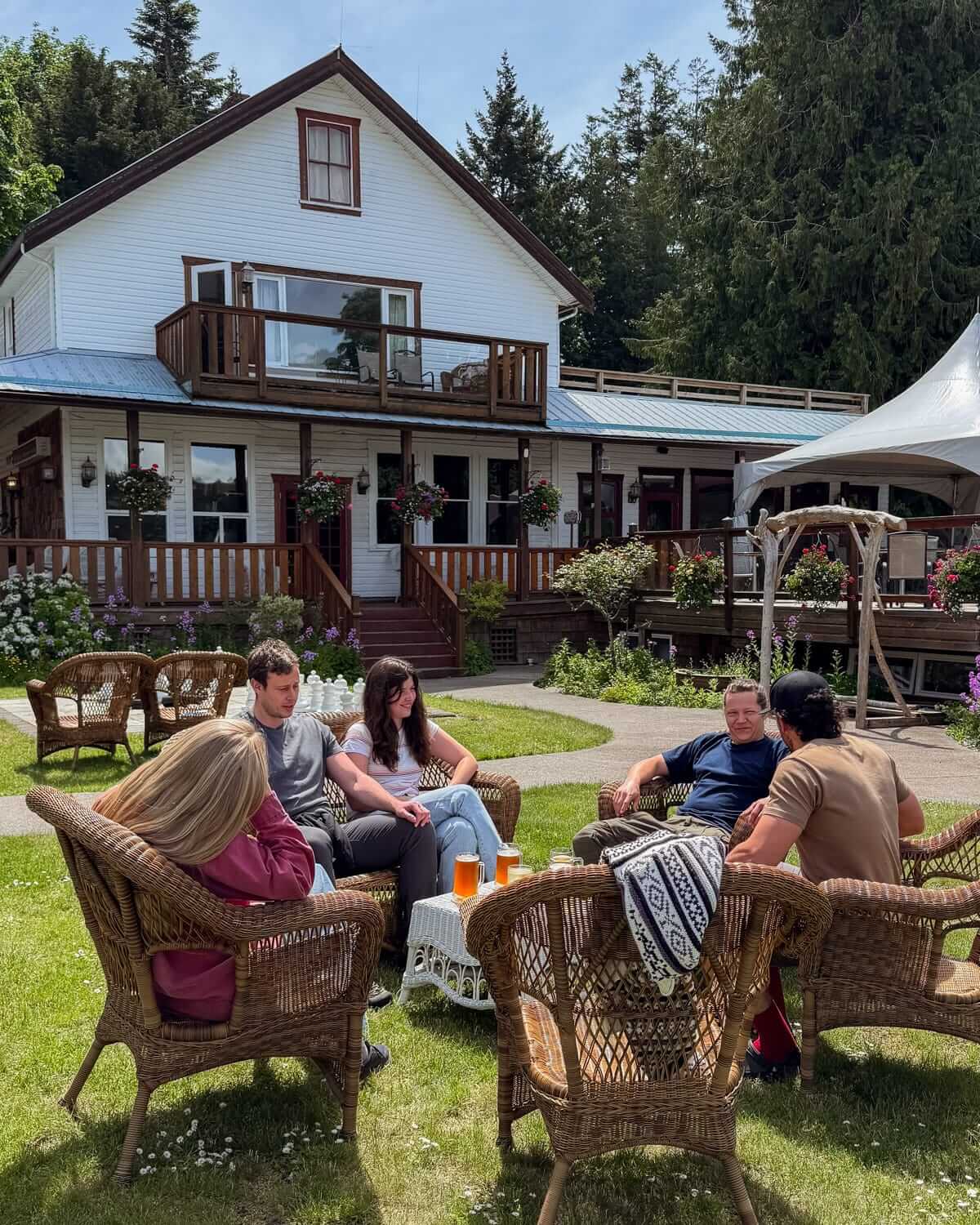 A group of friends sit in wicker chairs on the lawn of Heriot Bay Inn, enjoying drinks in the sunshine with a white heritage-style building in the background.