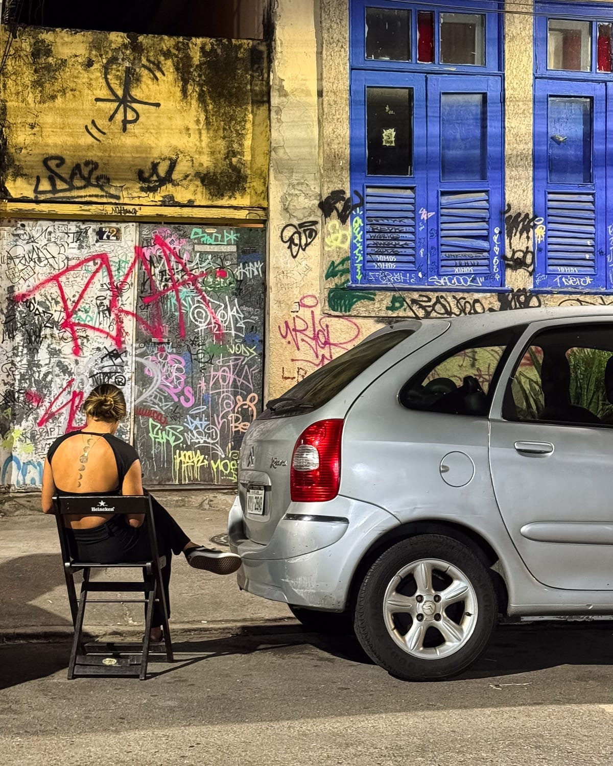 A girl sitting in a chair beside a parked car and graffiti in the lively Botafogo neighbourhood.