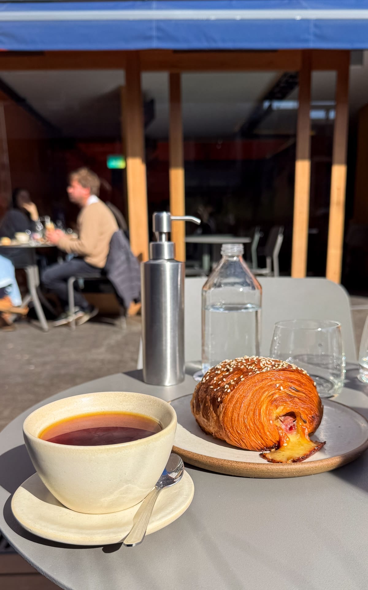 Outdoor table at Anchoita Panaderia with a cup of black coffee and a ham-and-cheese pastry, sunlight streaming across.