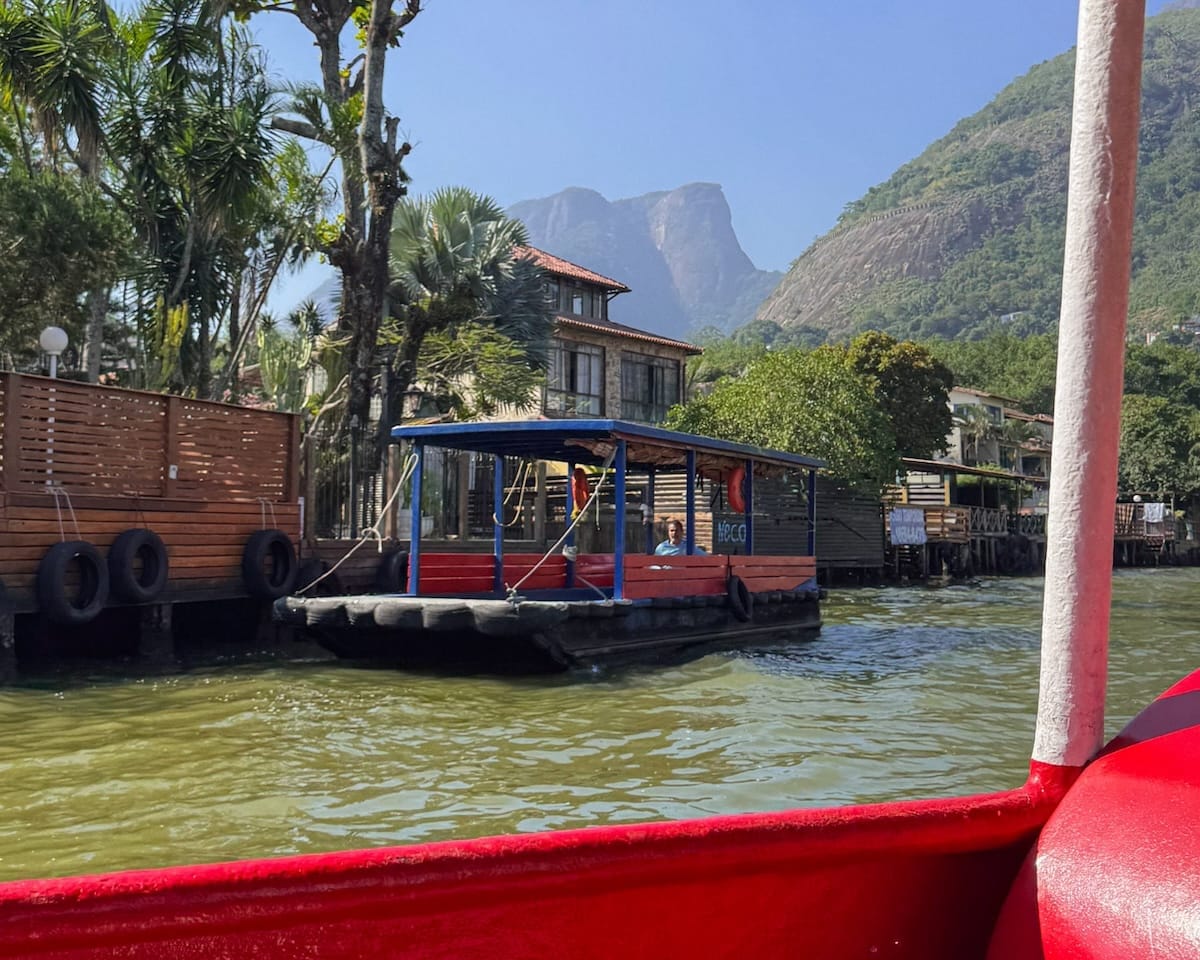 A small blue-and-red boat anchored along the wooden waterfront houses of Barra da Tijuca Lagoon, with lush palm trees and dramatic green mountains in the background.