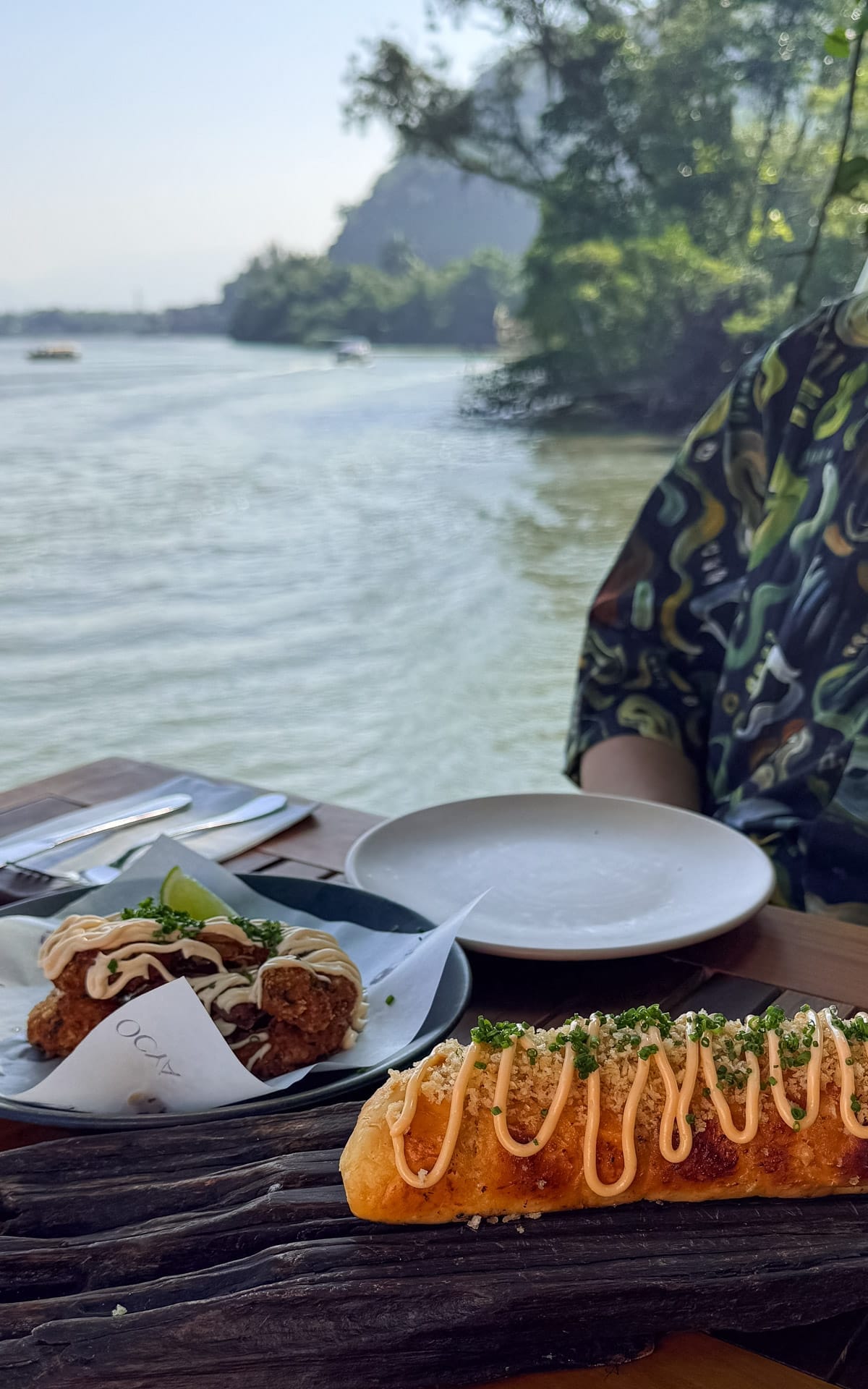  A close-up of an elegant lagoon-side table at Ocya Ilha featuring a shrimp bread appetizer drizzled with sauce and a plate of breaded shrimp bites, with calm lagoon waters and forested hills beyond.