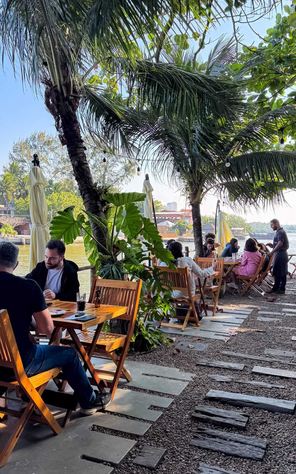 Guests dining at wooden tables shaded by palm trees at Ocya Ilha restaurant, with views of the tranquil lagoon.