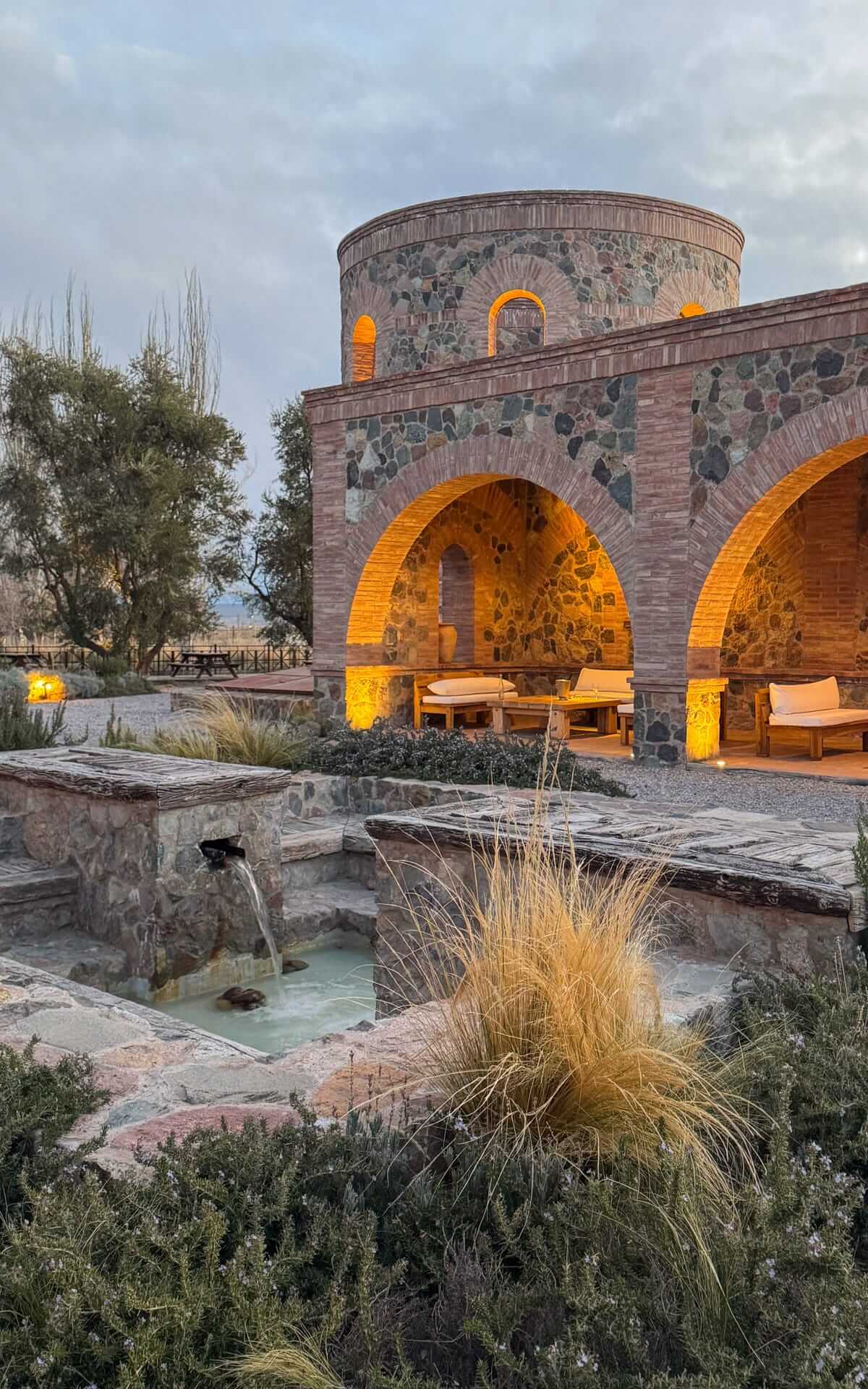 At Angelica restaurant at Catena Zapata, a rustic stone building with arched passageways glows warmly at dusk, with lounge chairs tucked beneath and a trickling fountain in front.