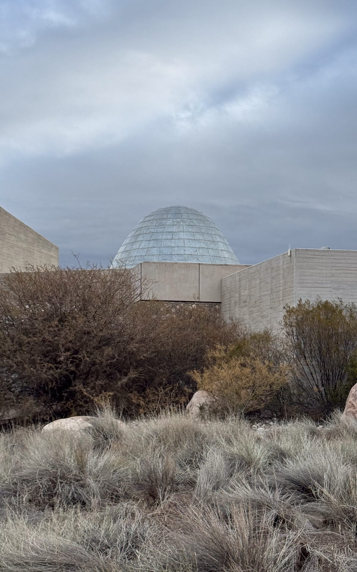 The modern dome-topped Zuccardi winery peeks over concrete walls and desert shrubs under a moody grey sky.