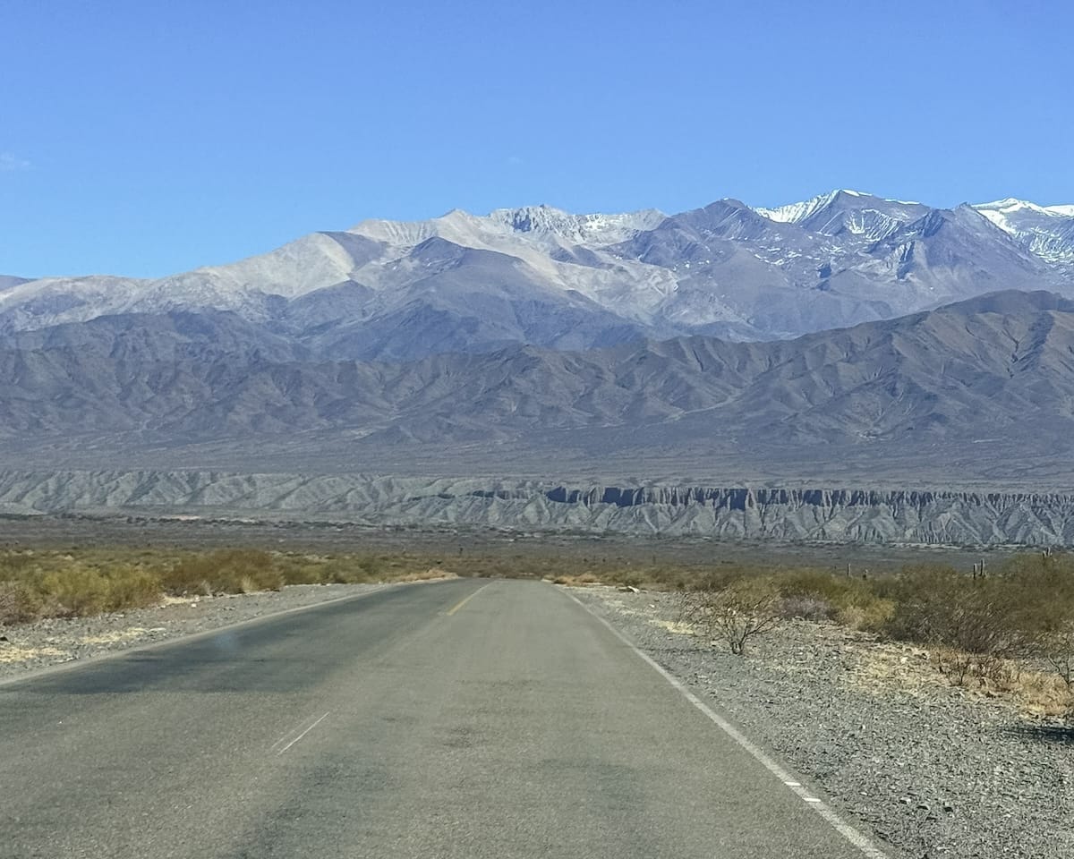 Empty paved road stretching towards layered desert mountains and the distant Andes.