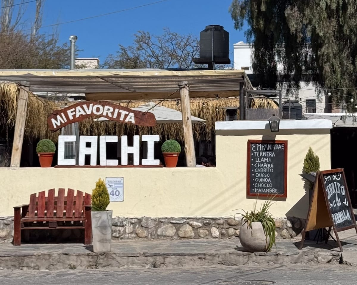 Sign reading “Mi Favorita Cachi” outside a rustic restaurant with an empanada menu and wooden bench in Cachi, Argentina.