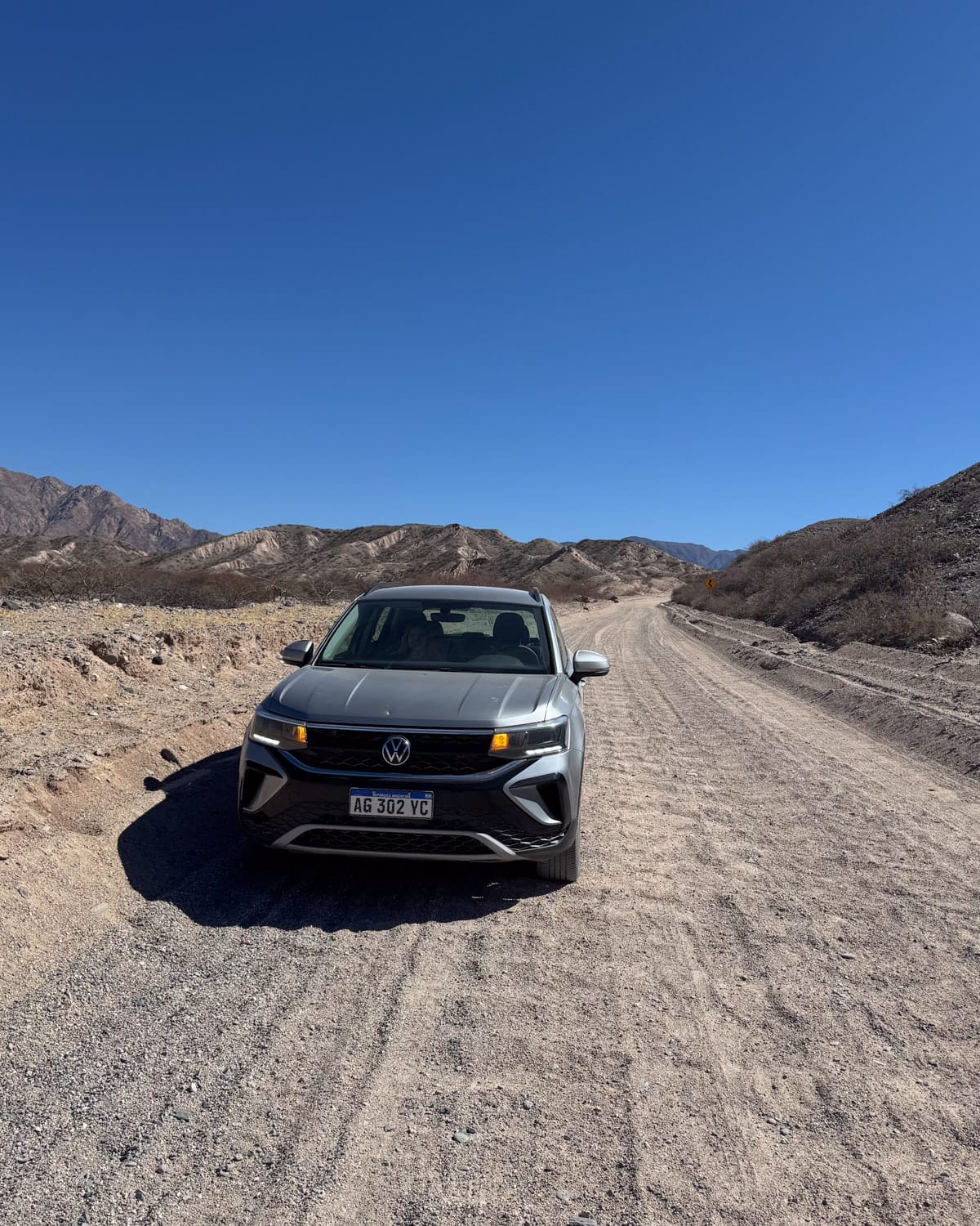 Silver Volkswagen parked on a gravel desert road surrounded by barren hills under a clear blue sky in Salta.