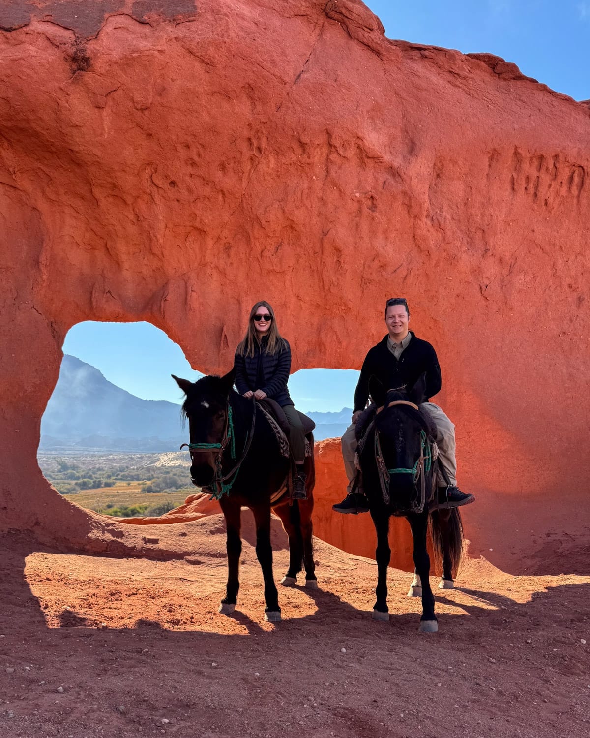 Cec and Ari on horseback beneath a red rock arch with sweeping desert and mountain views near Cafayate.