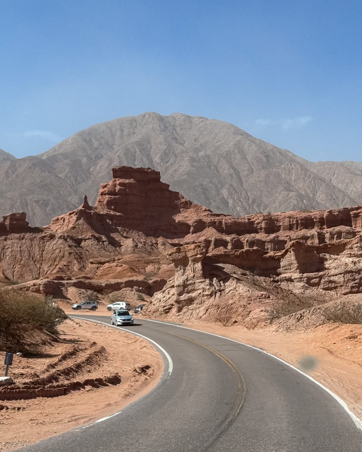 Cars winding through a paved road surrounded by red rock formations and desert mountains in northern Argentina.