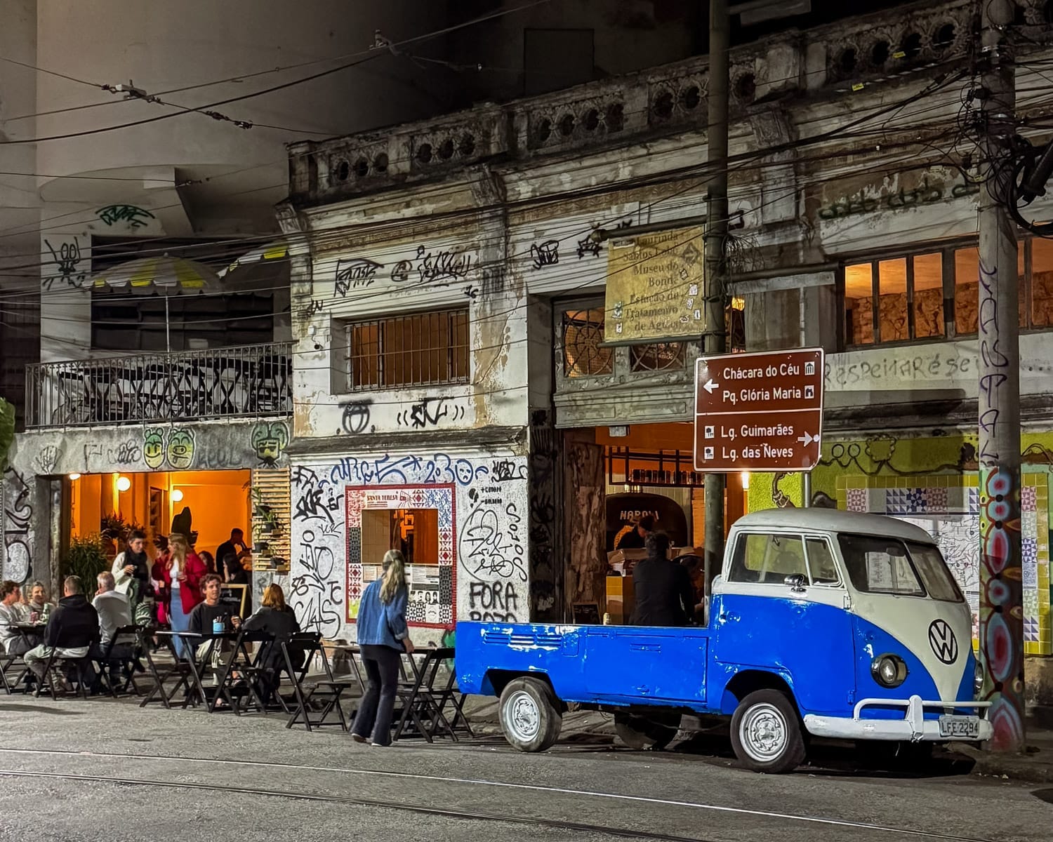 Nighttime street scene in Santa Teresa with people sitting at outdoor tables, graffiti-covered buildings, and a blue vintage Volkswagen truck parked out front.