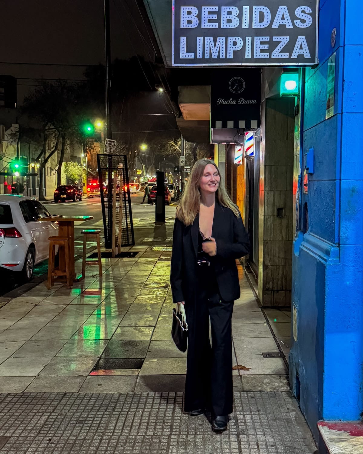 Cec in black outfit walks along a sidewalk at night under a bright “Bebidas Limpieza” sign in Buenos Aires.