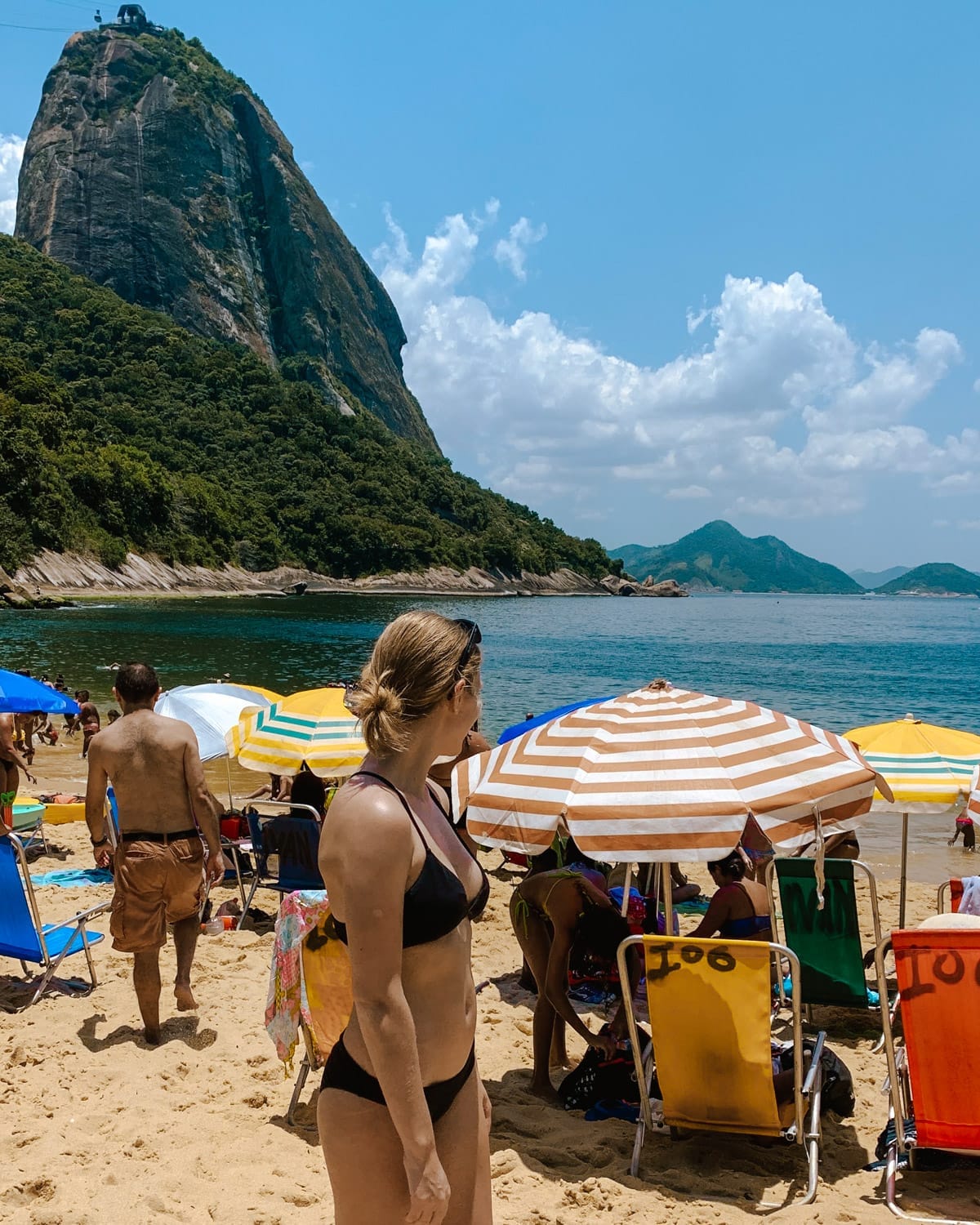 Cec standing on Praia Vermelha with striped beach umbrellas, calm blue water, and Sugarloaf Mountain towering in the background.