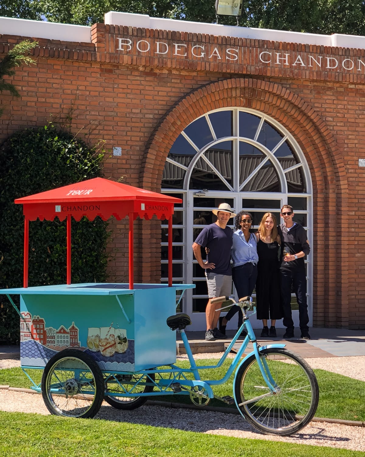 Cec, Ari and Friends outside Bodegas Chandon in Mendoza beside a blue cart with red canopy on a sunny day.