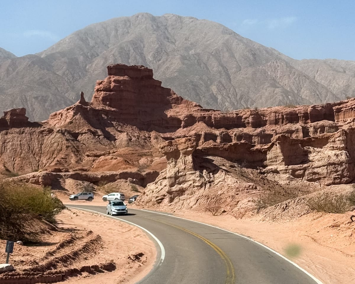 Cars winding through a paved road surrounded by red rock formations and desert mountains in northern Argentina.