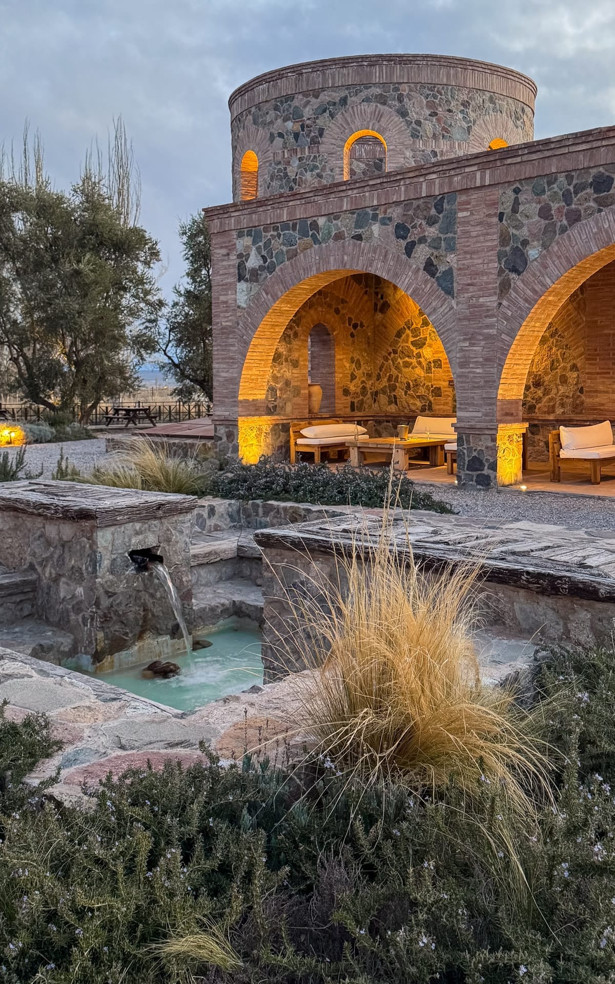 Stone courtyard and arched brick building at Angélica Bistro, softly lit at dusk with a small fountain in front.