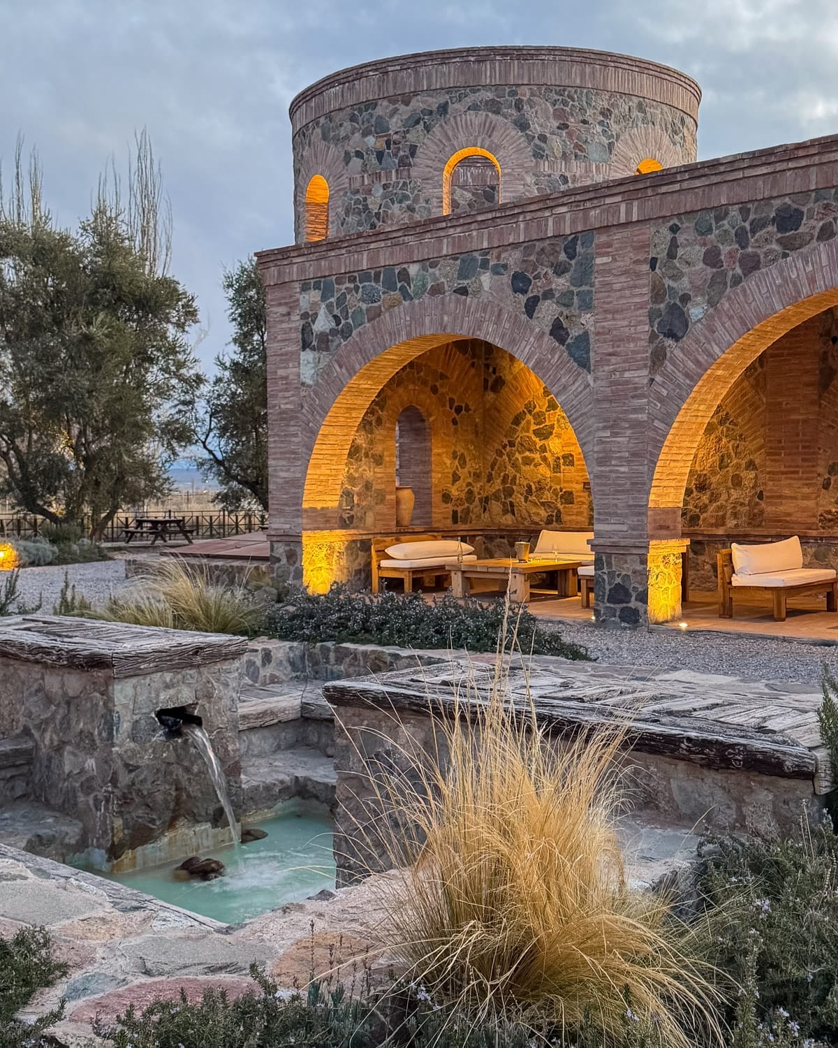Stone courtyard and arched brick building at Angélica Bistro, softly lit at dusk with a small fountain in front.