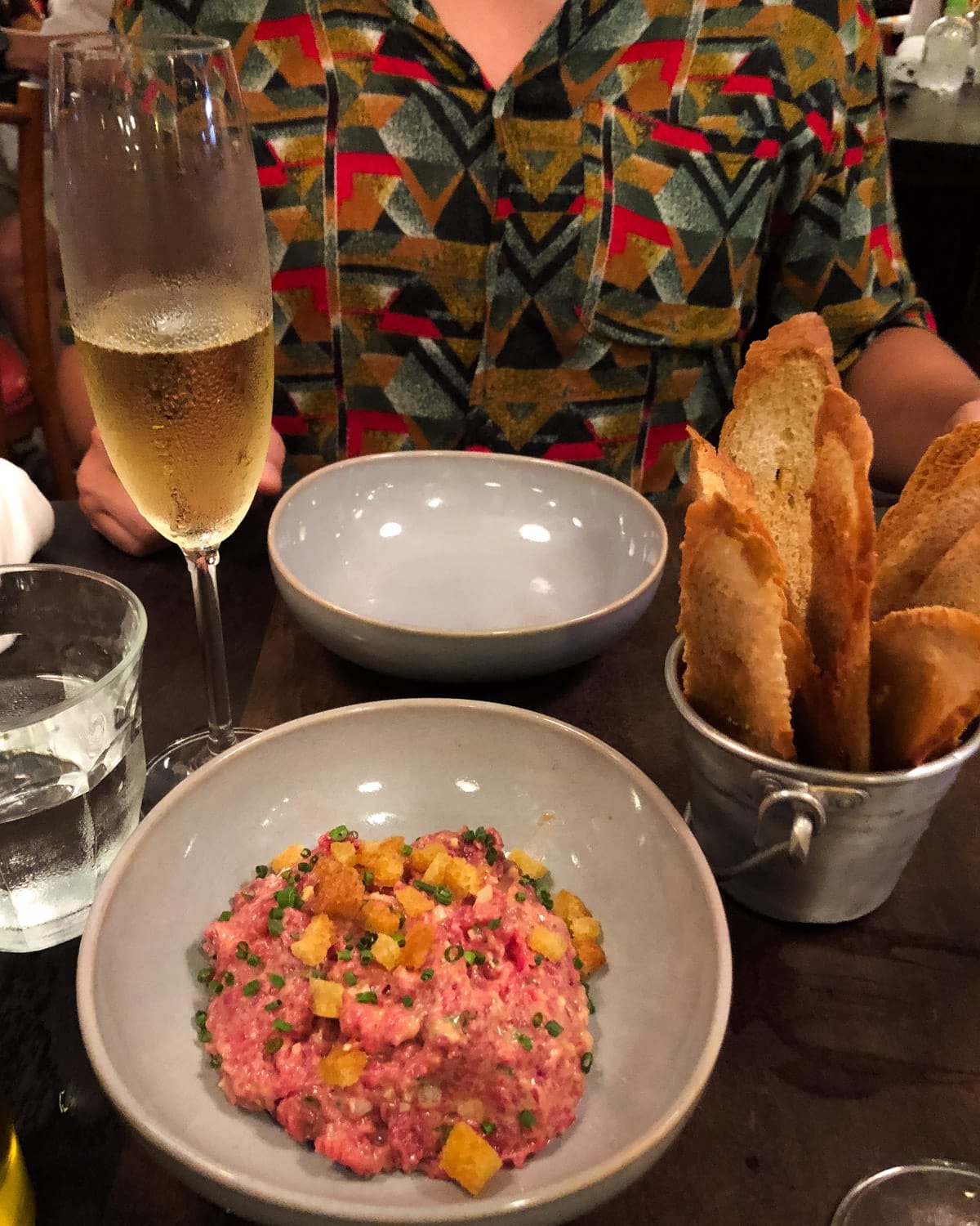 Beef tartare topped with chopped chives and golden croutons in a gray ceramic bowl and glass of Brazilian sparklingat wine a CT Boucherie in Rio.