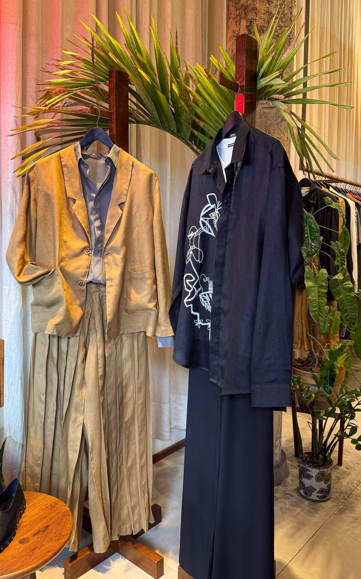 Display of contemporary Brazilian designer clothing at Handred, featuring neutral-toned linen pieces and a black embroidered shirt beside tropical plants.
