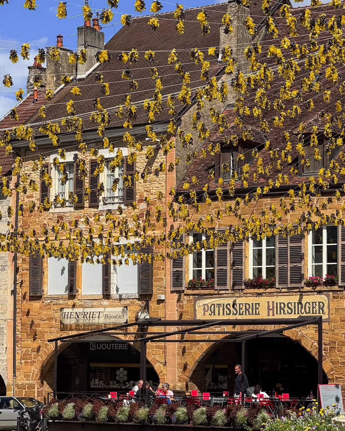 Café tables set in Arbois town square beneath hanging decorations, with stone buildings and the Édouard Hirsinger chocolatier storefront behind.