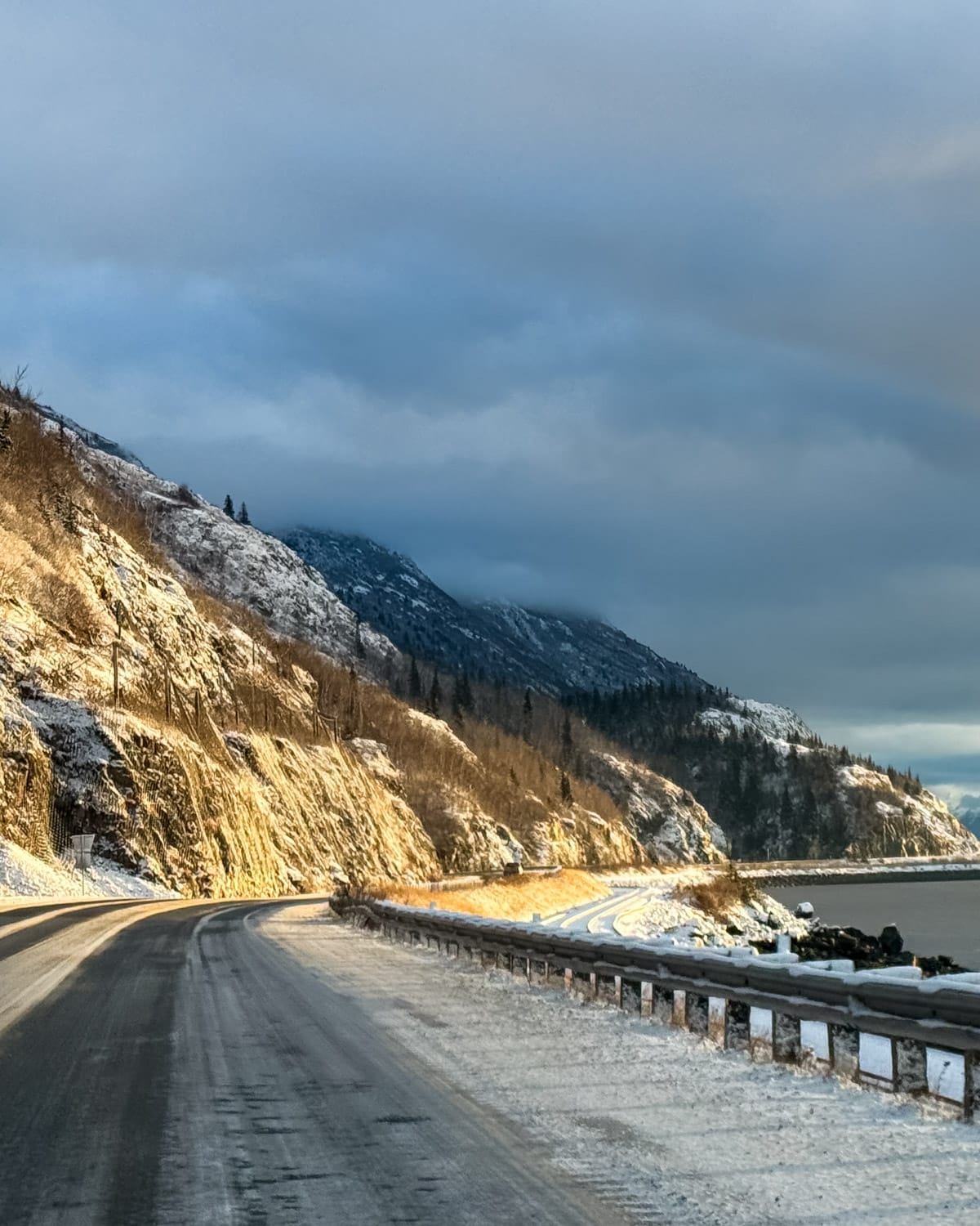 Snow-covered coastal highway curving along Turnagain Arm near Anchorage, with icy guardrails, forested cliffs, and distant mountains under a dramatic winter sky.